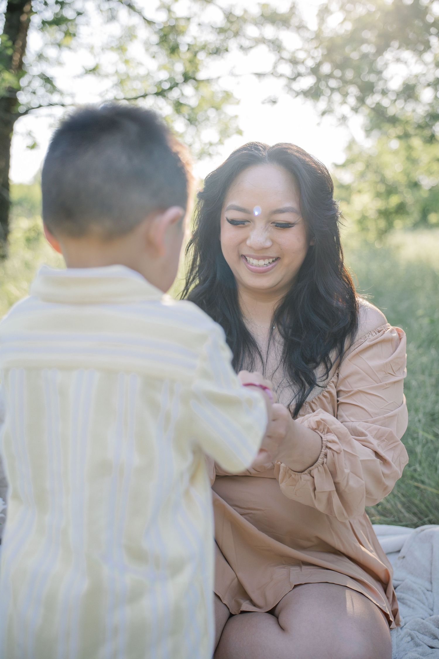 A smiling woman in a beige dress holds hands with a young boy outdoors in a sunlit green field.