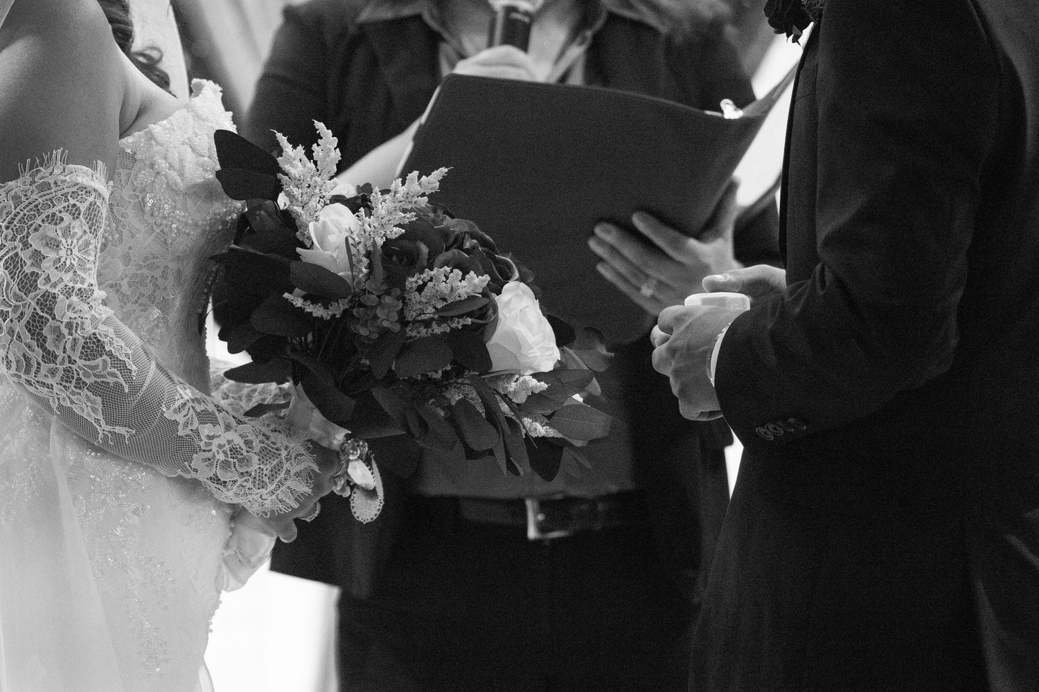 Bride holding dark floral bouquet during wedding ceremony while officiant reads vows, black and white.