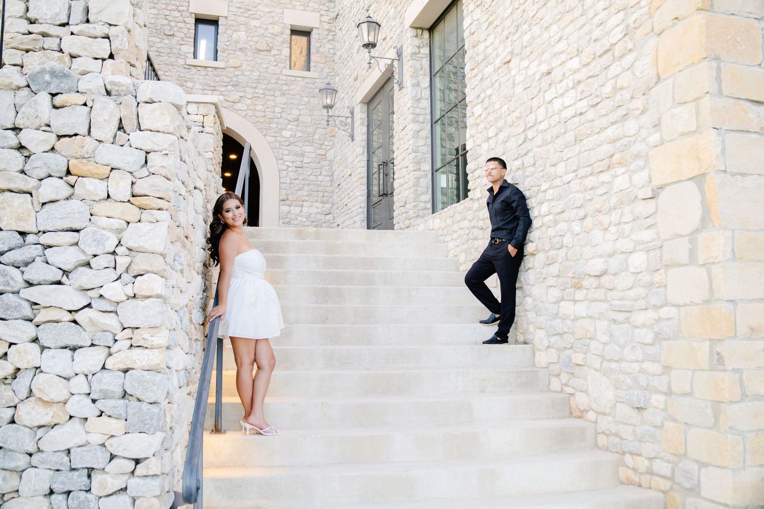 Couple in elegant attire posing on stone steps outside a rustic stone building.