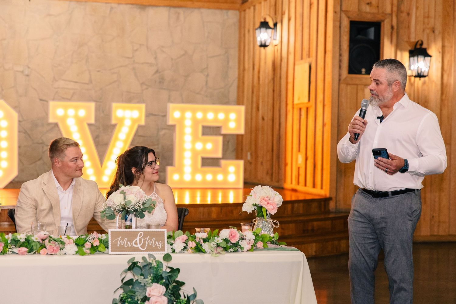 Bride and groom listen at the head table as a man gives a wedding toast in front of large illuminated LOVE marquee letters.