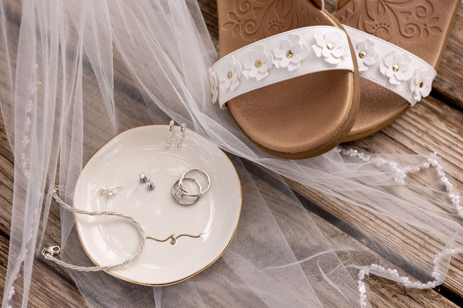 Wedding rings and jewelry rest on a small white dish beside floral sandals and a sheer veil on rustic wood.