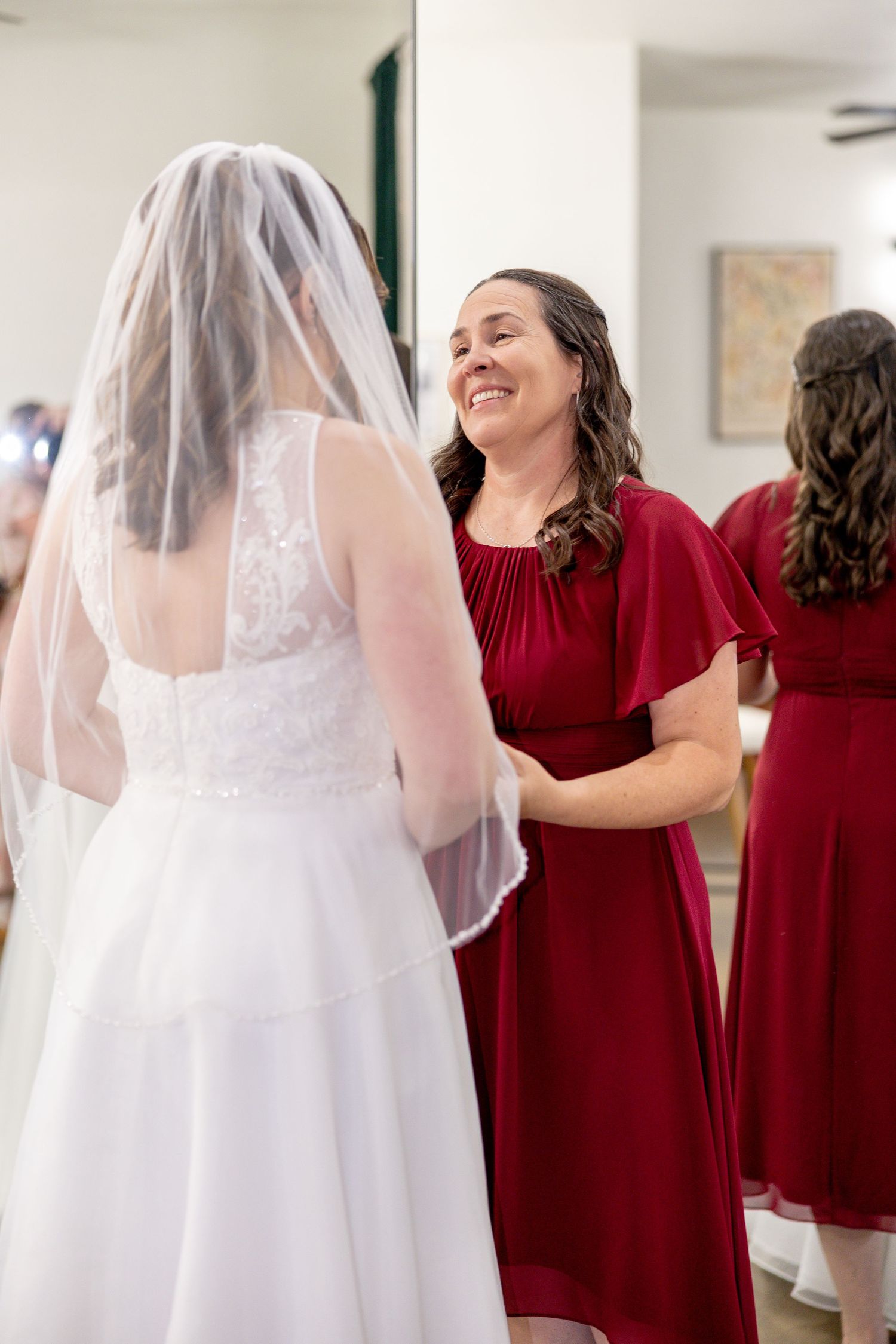 Smiling bridesmaid in a red dress talks with a bride in a white lace gown and long veil before the wedding ceremony.