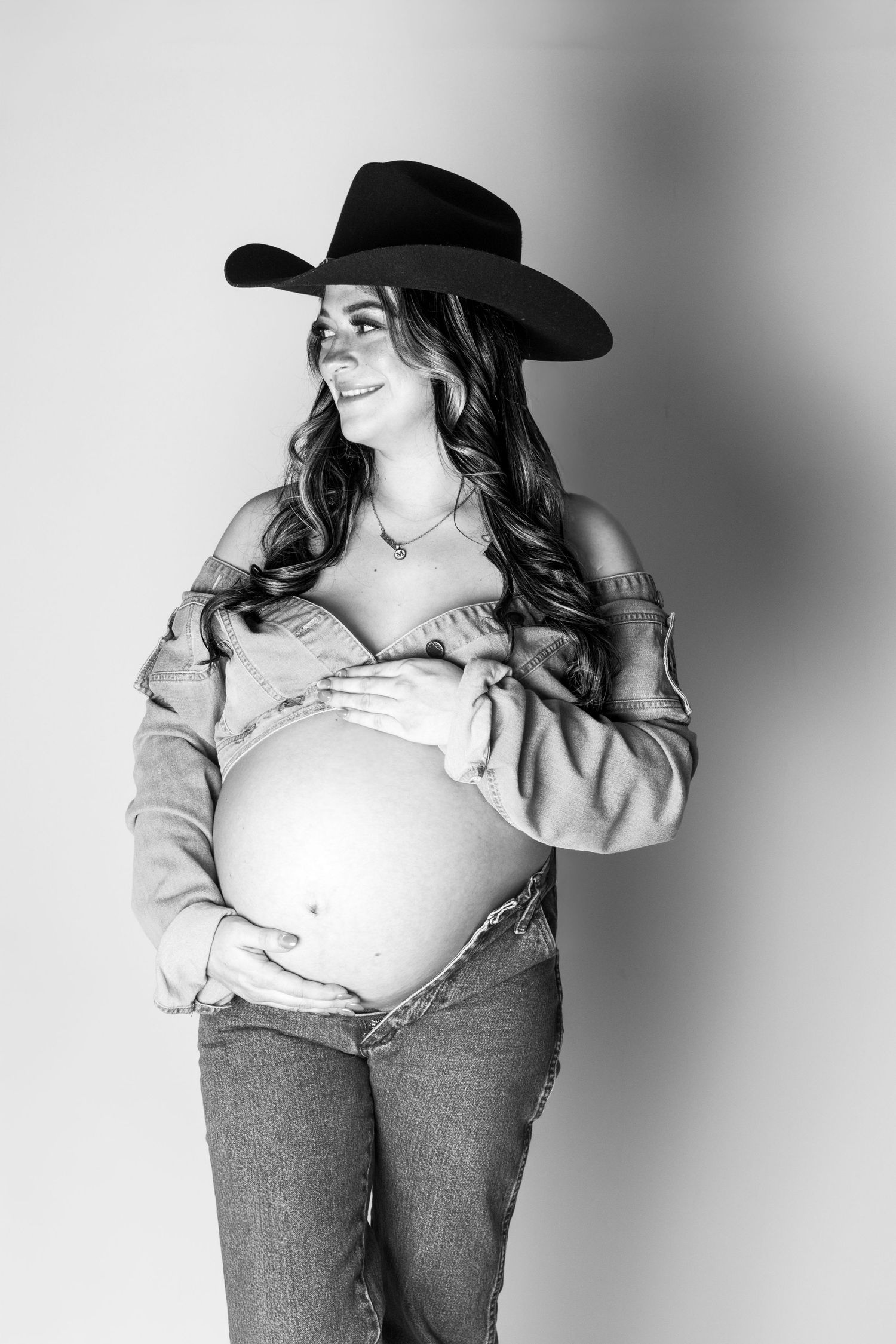 Black and white photo of a smiling pregnant woman in a cowboy hat and jeans cradling her baby bump.