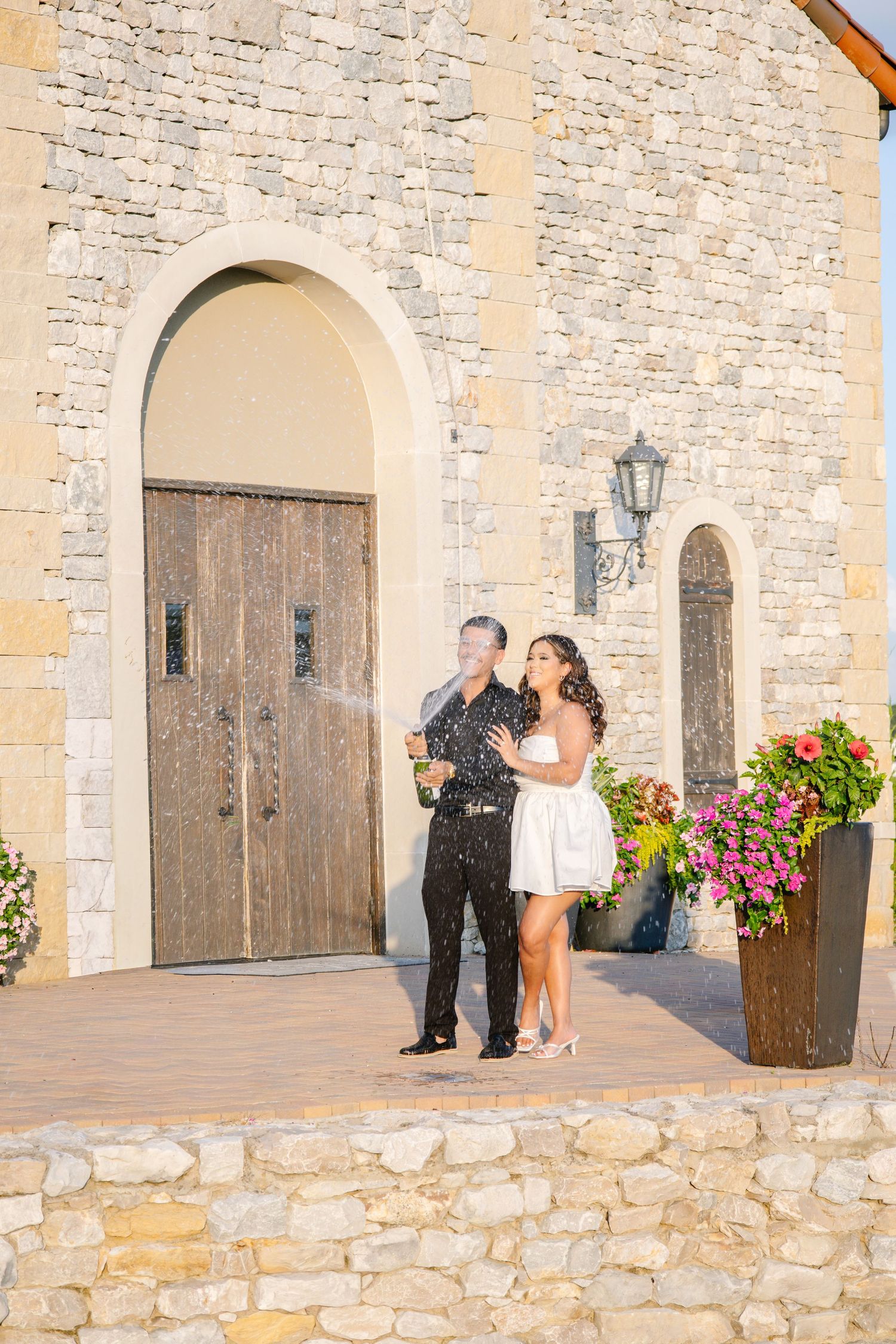 Couple celebrating with champagne outside a stone building with arched wooden door and floral decor.