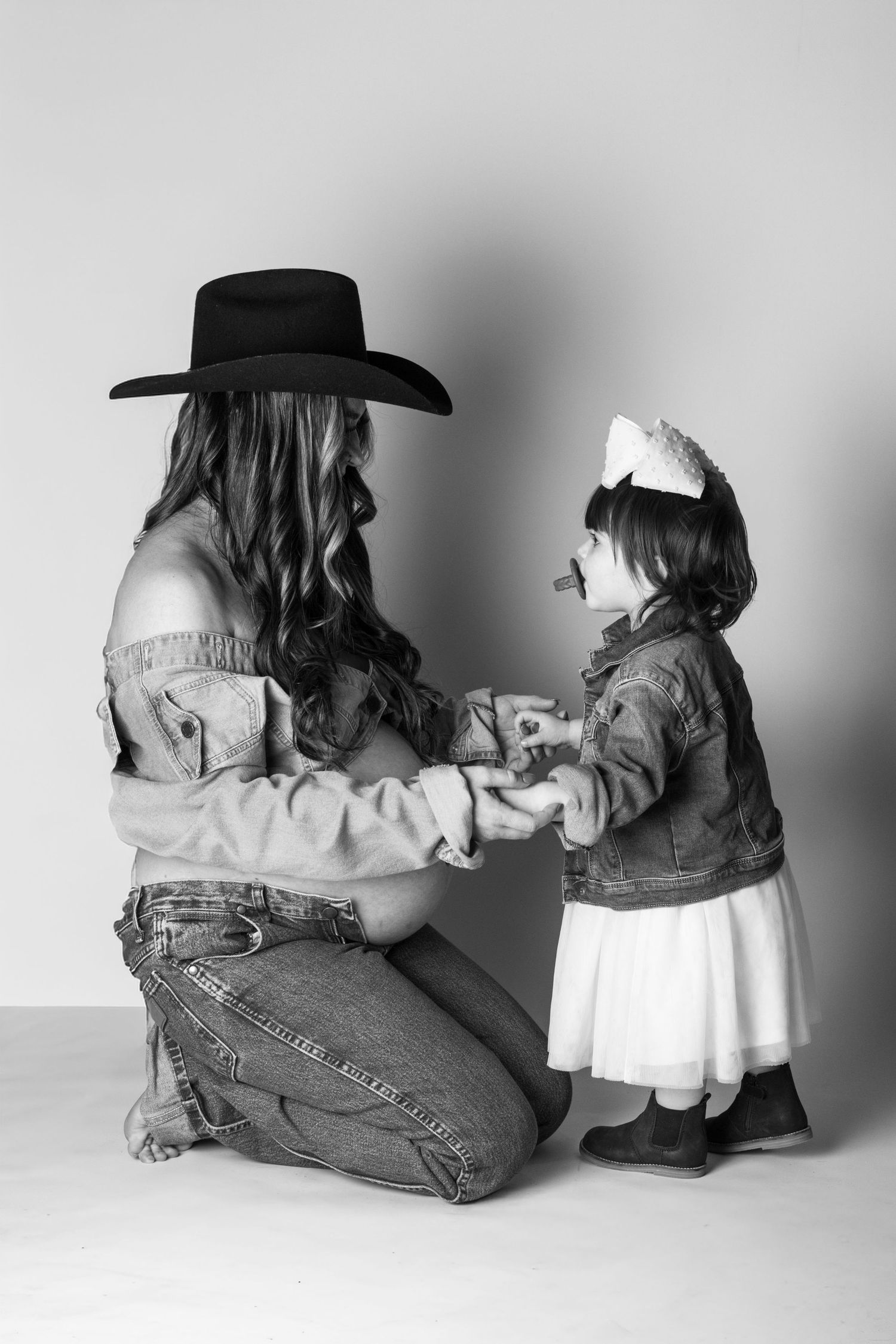 Black and white photo of mother kneeling in cowboy hat holding hands with toddler daughter in denim jacket and white skirt.