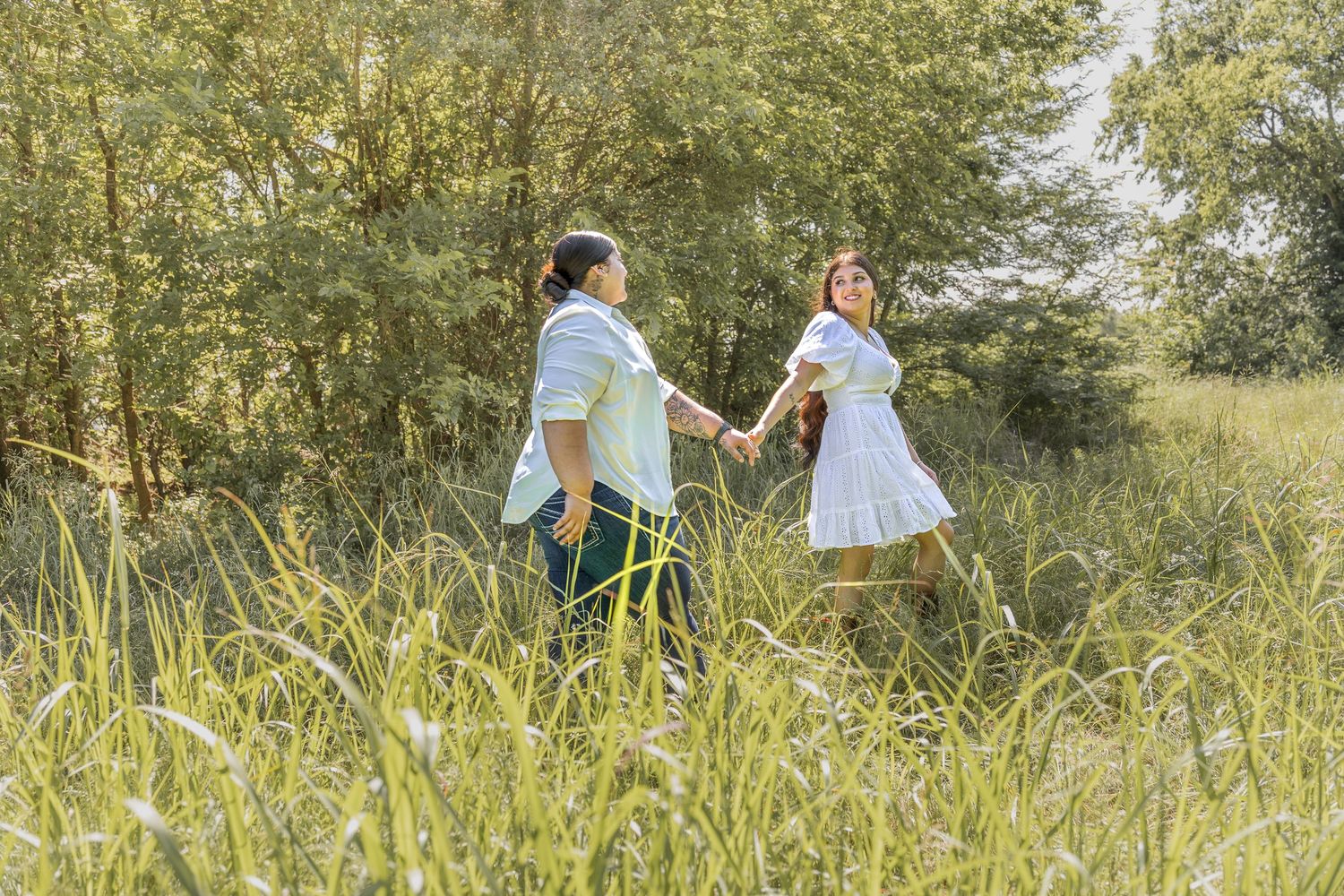 Two women walk hand in hand through a lush green meadow on a bright sunny day.