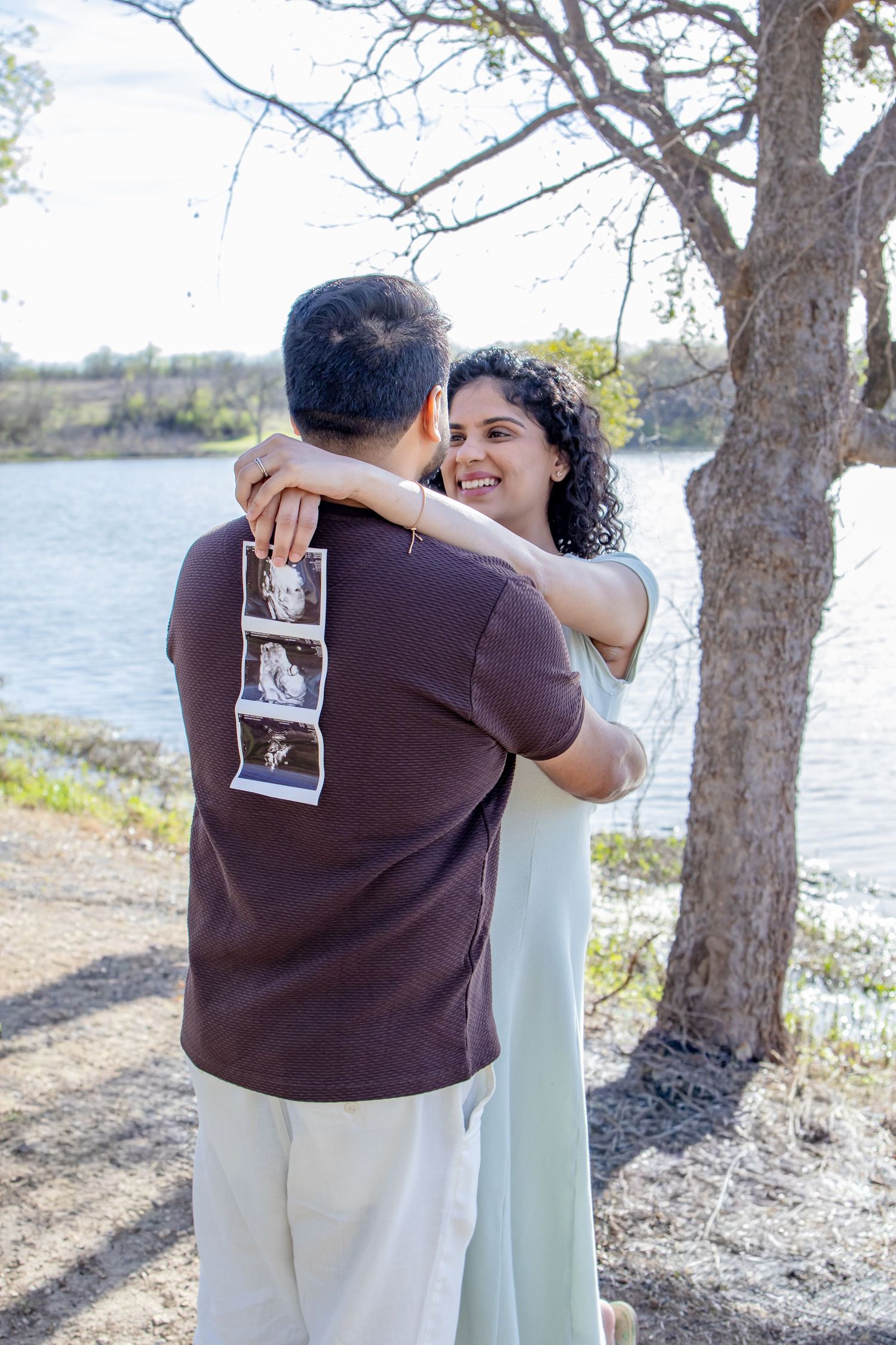 Smiling pregnant woman hugging her partner outdoors near a lake, holding ultrasound photos behind his back.