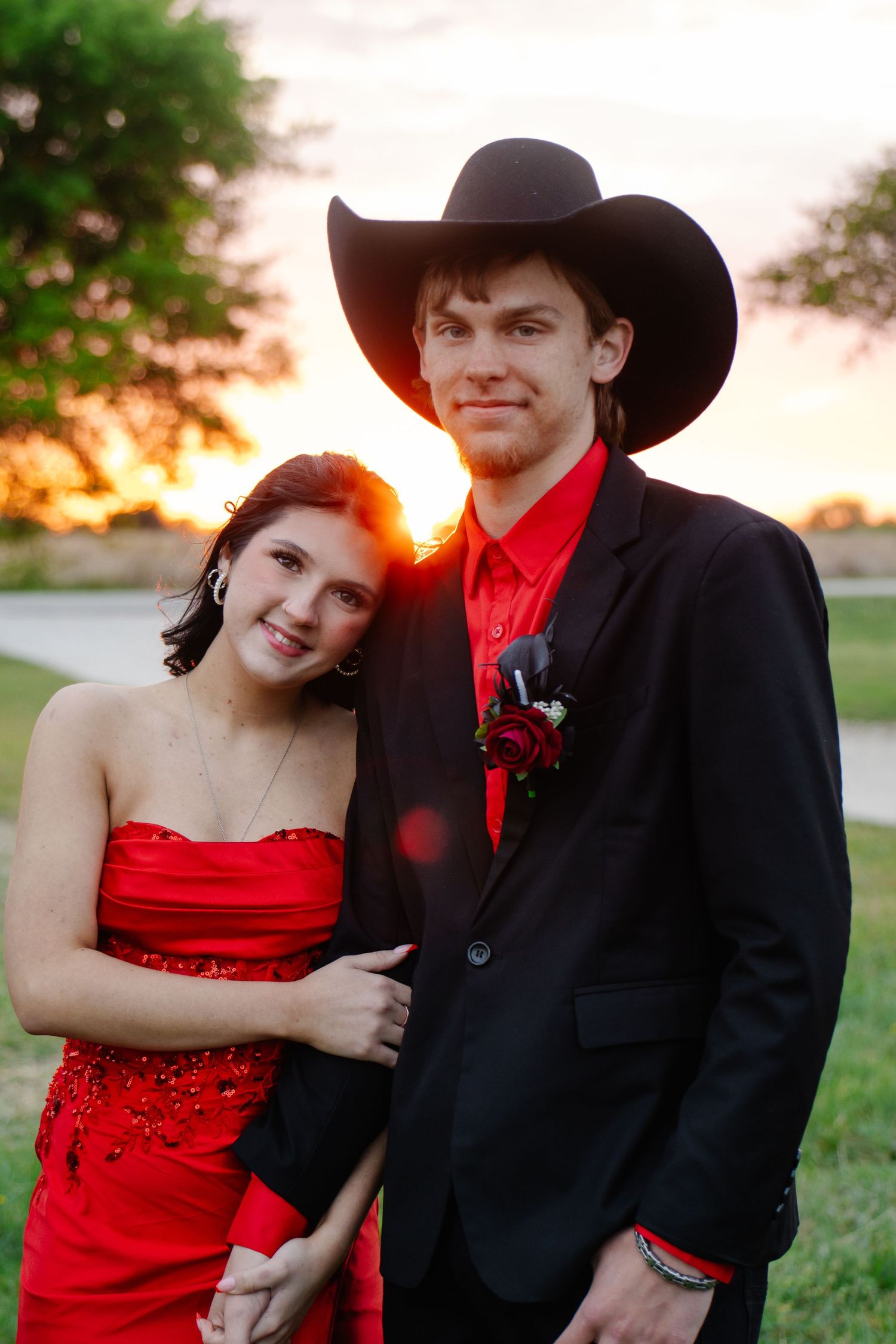Smiling prom couple at golden hour, girl in red dress leaning on boy in black suit and cowboy hat.