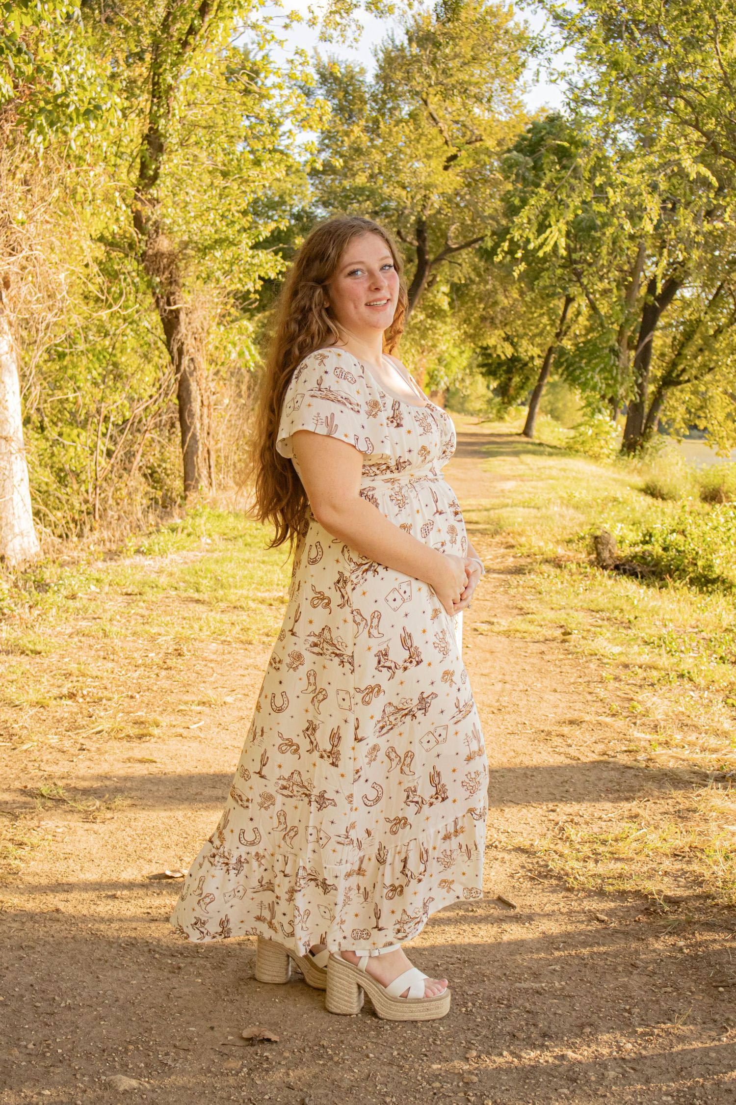 Pregnant woman in a floral maxi dress standing on a sunlit dirt path surrounded by golden autumn trees.