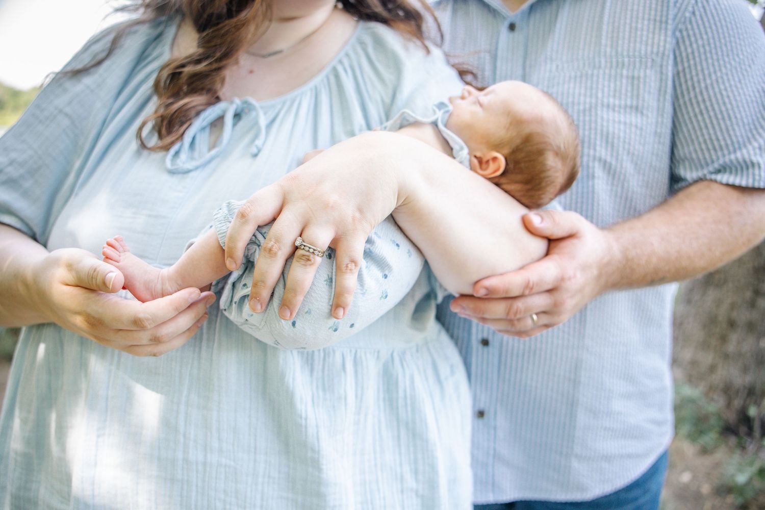 Parents cradling their newborn baby, mother wearing rings and a light blue dress, father in a striped shirt, outdoors.
