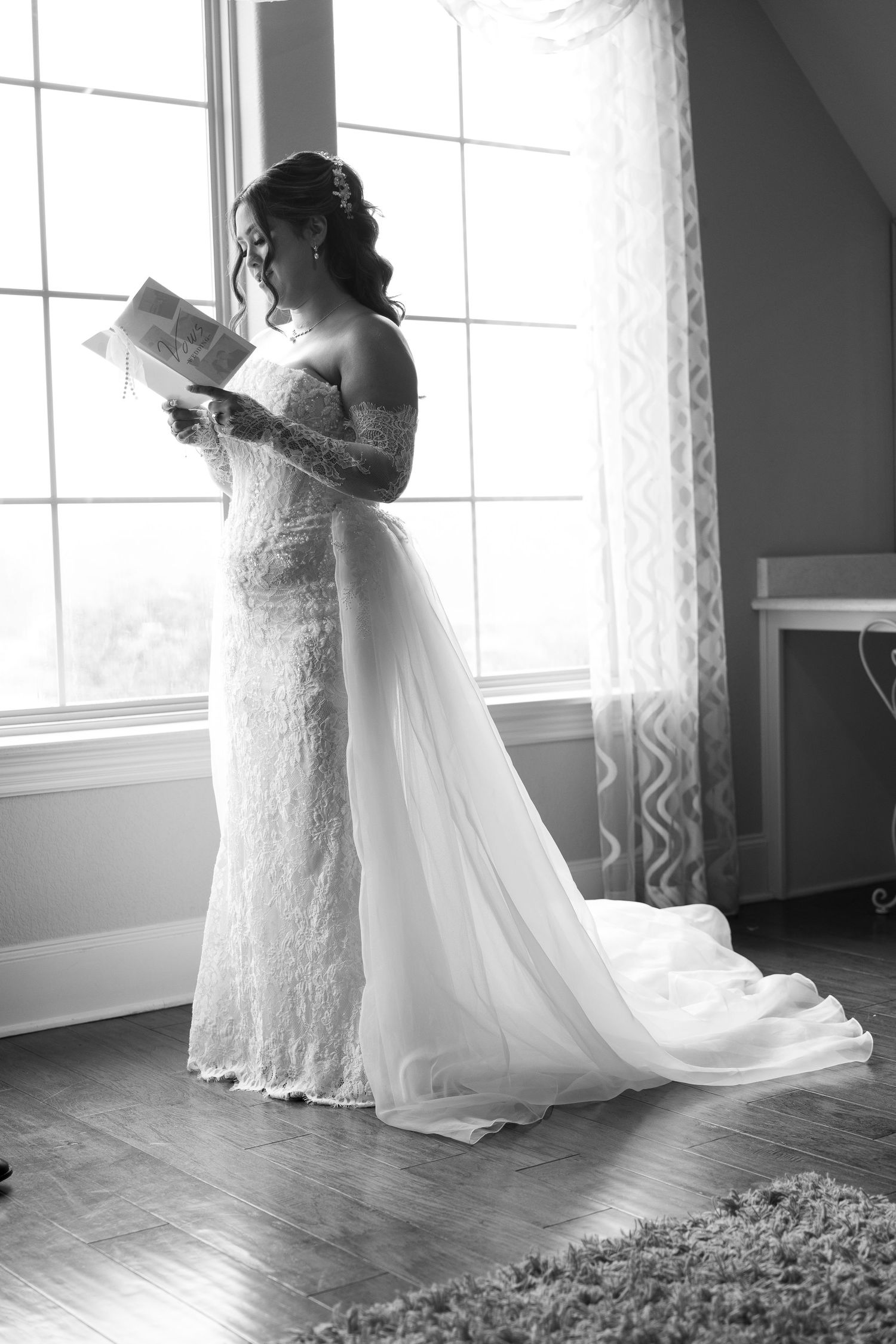 Black and white portrait of a bride in a lace gown reading a letter by a large window.