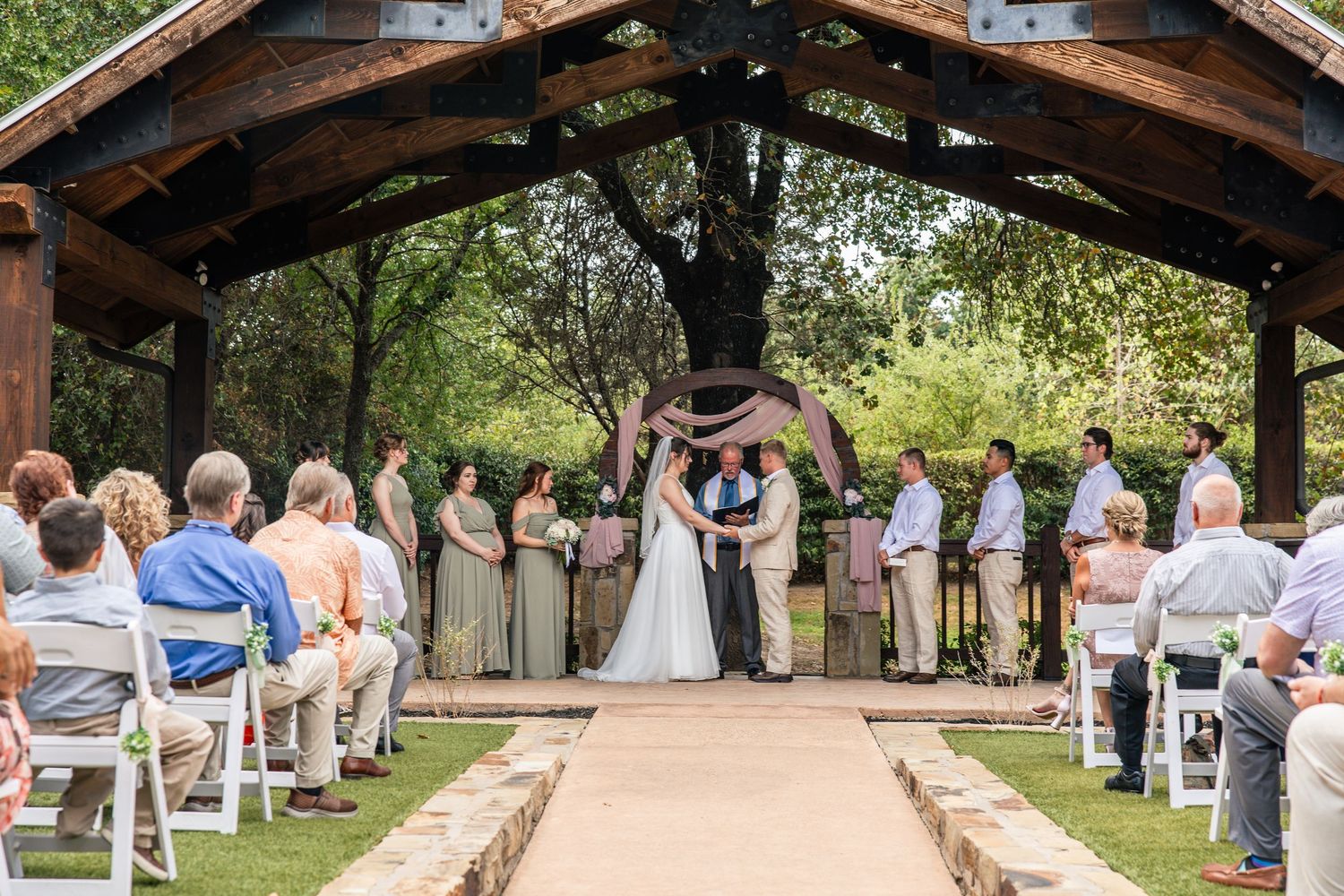 Bride and groom exchange vows under a rustic wooden pavilion arch while wedding guests watch from white chairs outdoors.