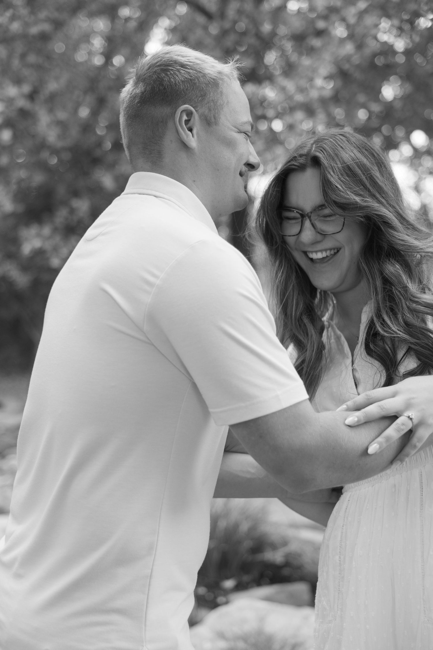 Happy couple laughing together outdoors, man in white shirt embracing smiling woman with glasses, black and white photo.