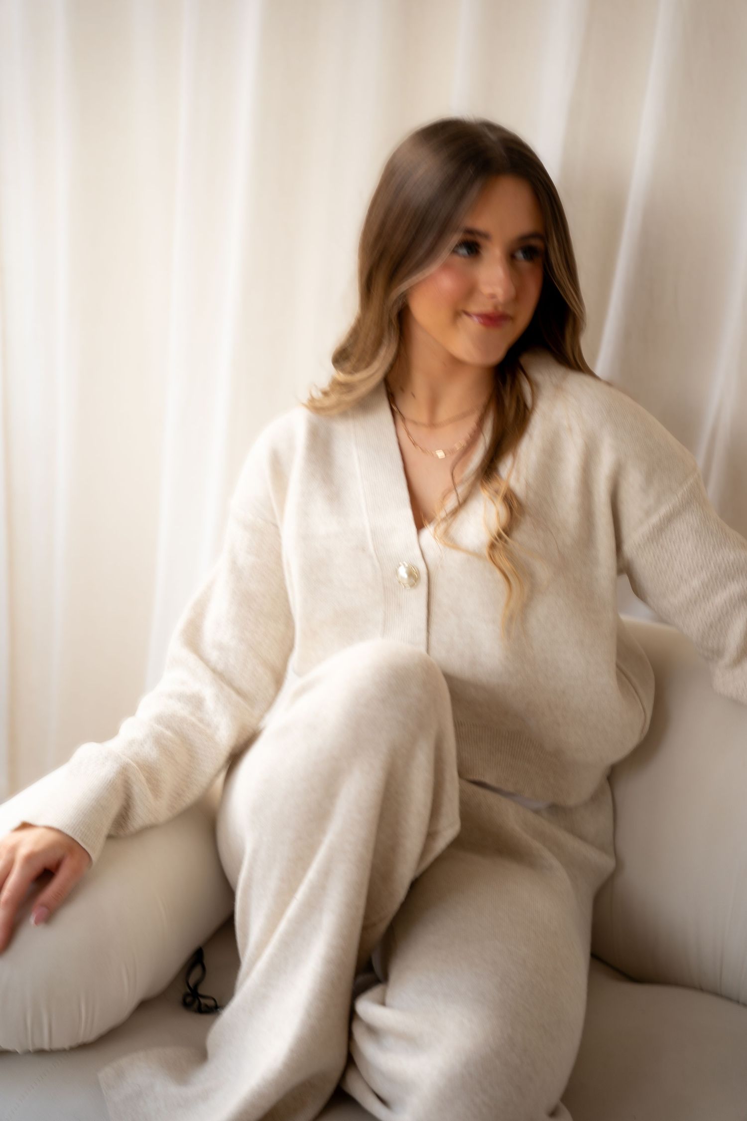 Blonde woman in beige knit cardigan and trousers sitting relaxed on white chair by curtains.