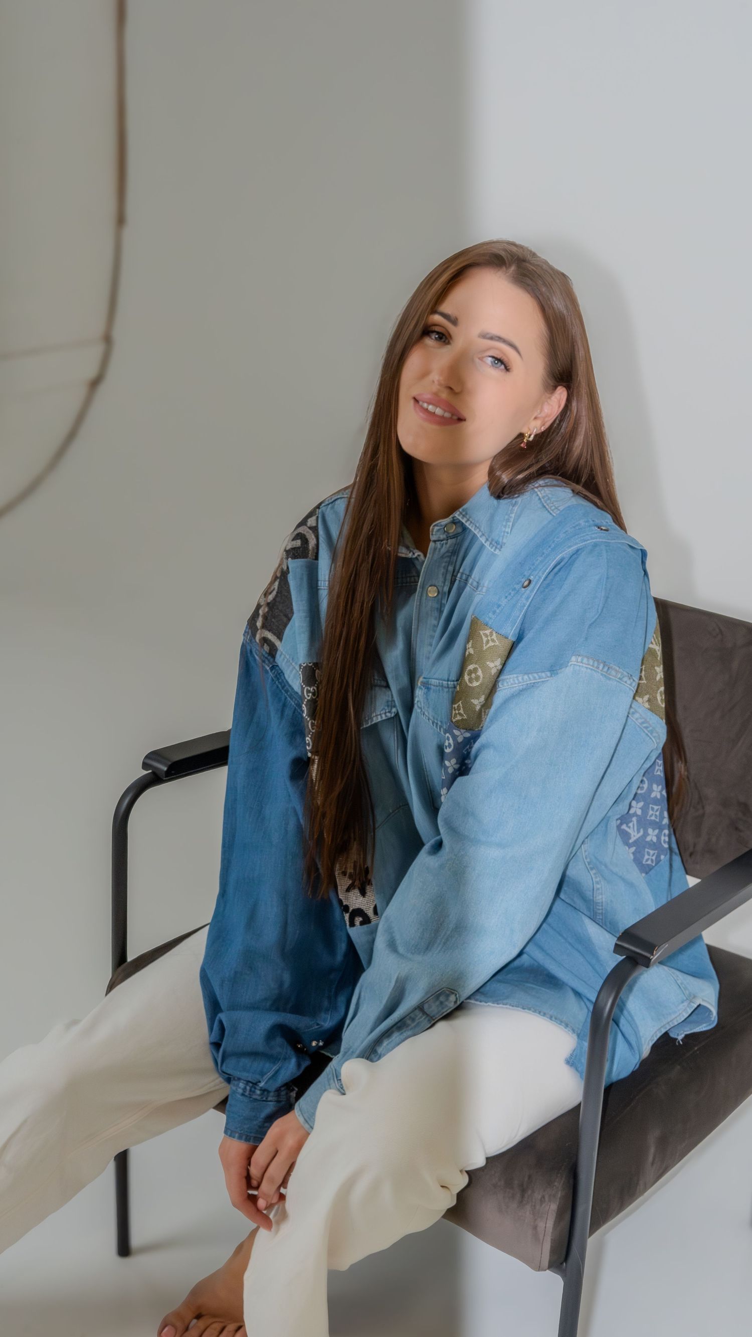 Brunette woman in patchwork denim jacket and white pants seated on metal chair in studio.