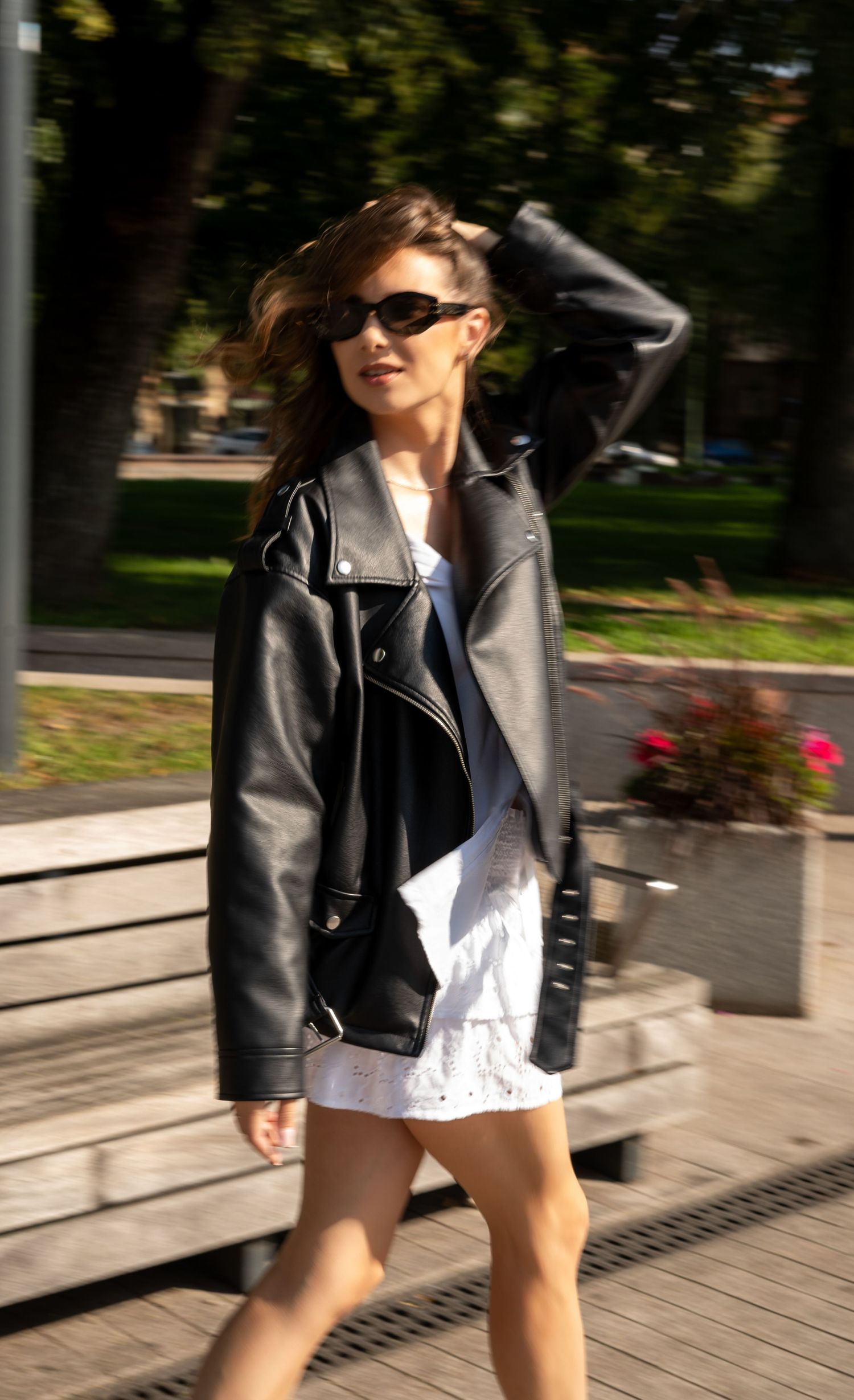 Stylish woman wearing black leather biker jacket and white dress posing outdoors in sunlight.