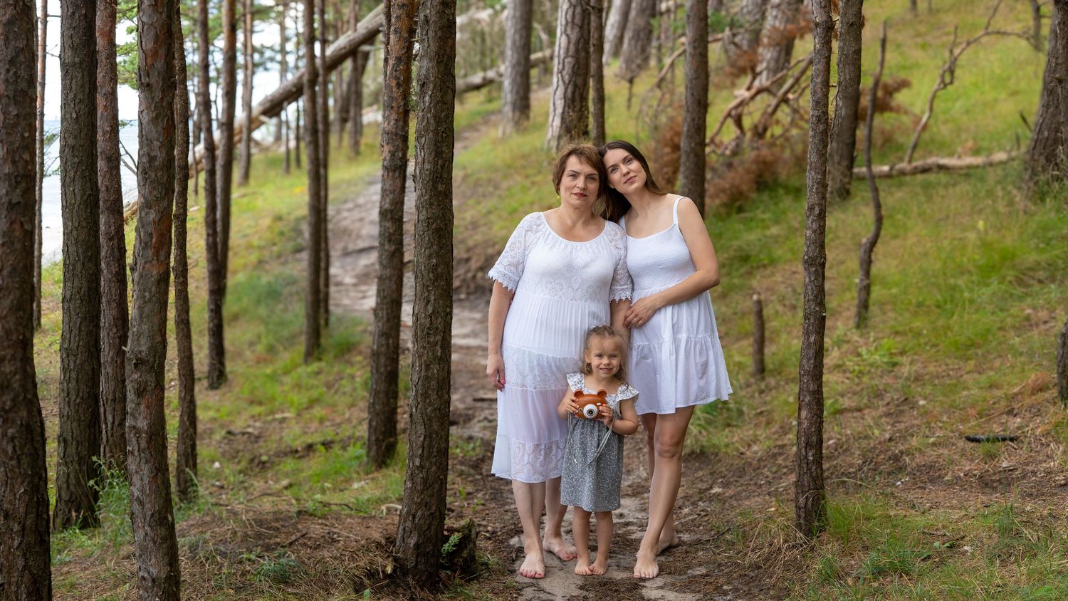 Three generations of women in white dresses stand together in a pine forest near water.