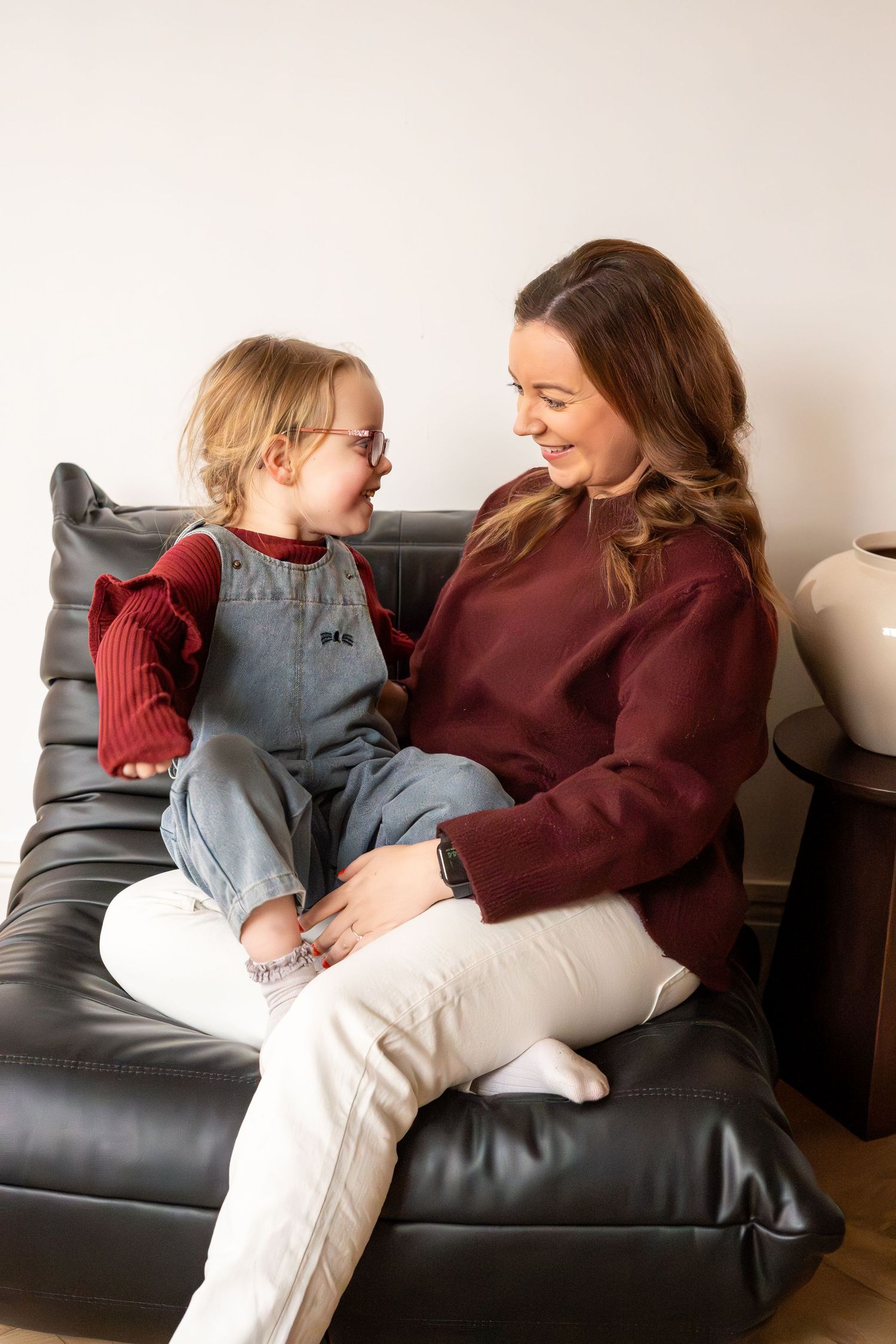 A smiling mother and young girl with glasses share a joyful moment sitting on a black leather chair.