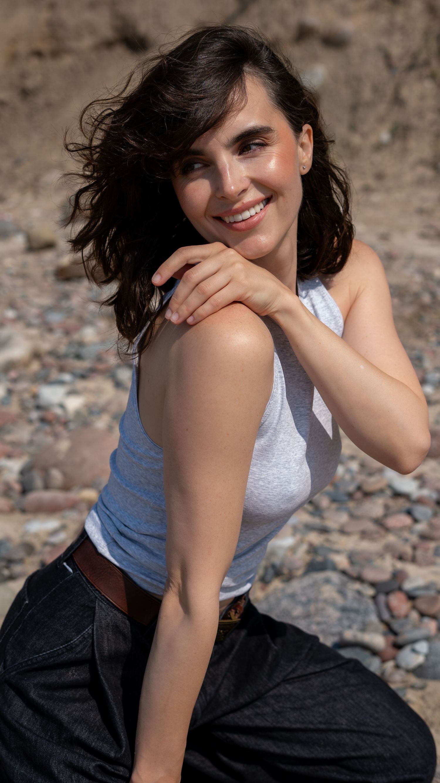 Smiling brunette woman in white sleeveless top and black jeans posing on rocky beach.