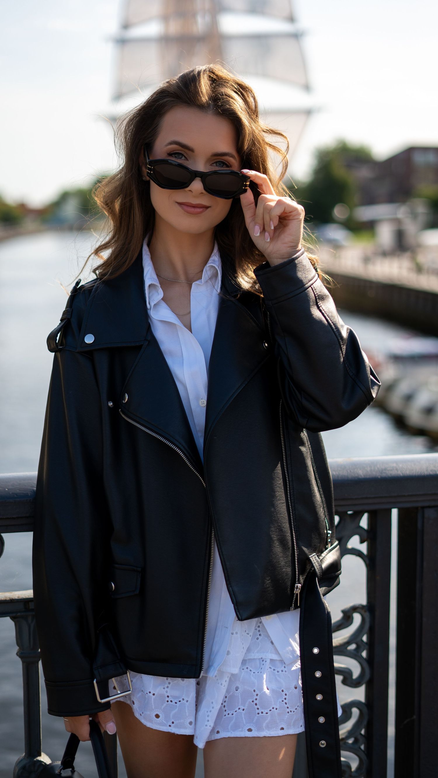 Stylish woman in black leather jacket and white dress holding sunglasses near waterfront on sunny day.