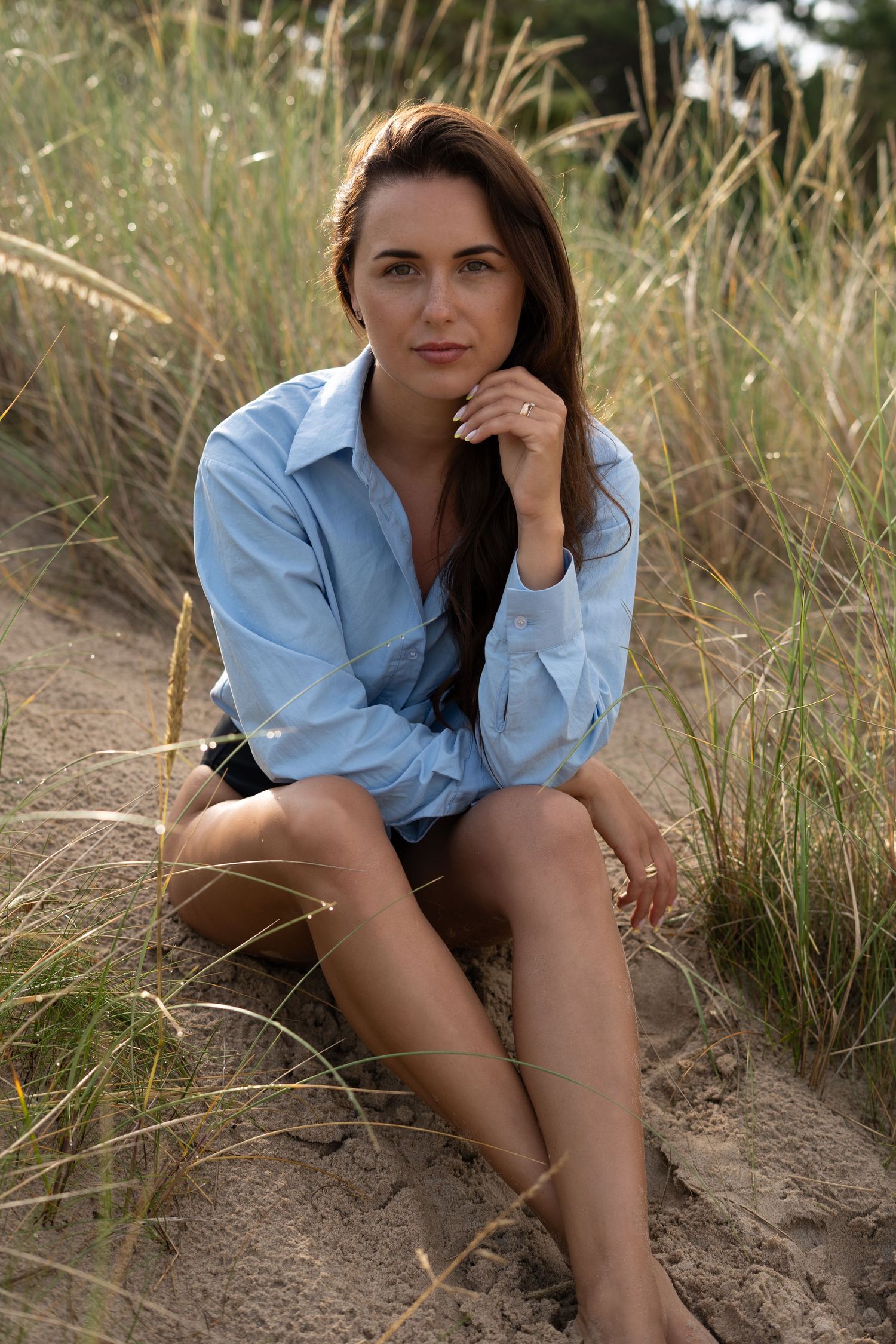 Brunette woman in blue oversized shirt sitting on sandy dunes with long grass in background.