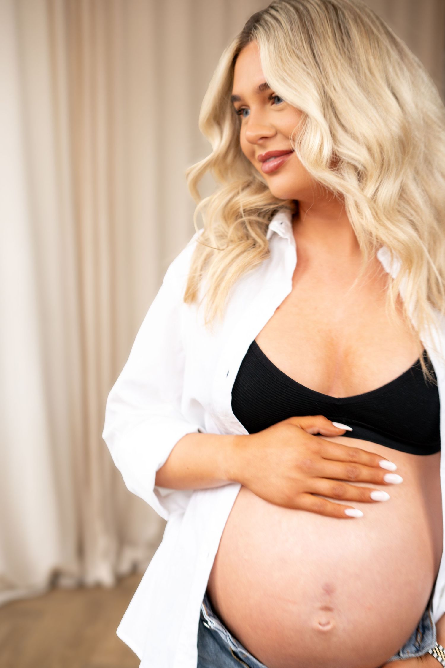Pregnant blonde woman in open white shirt and black bralette tenderly holding baby bump.