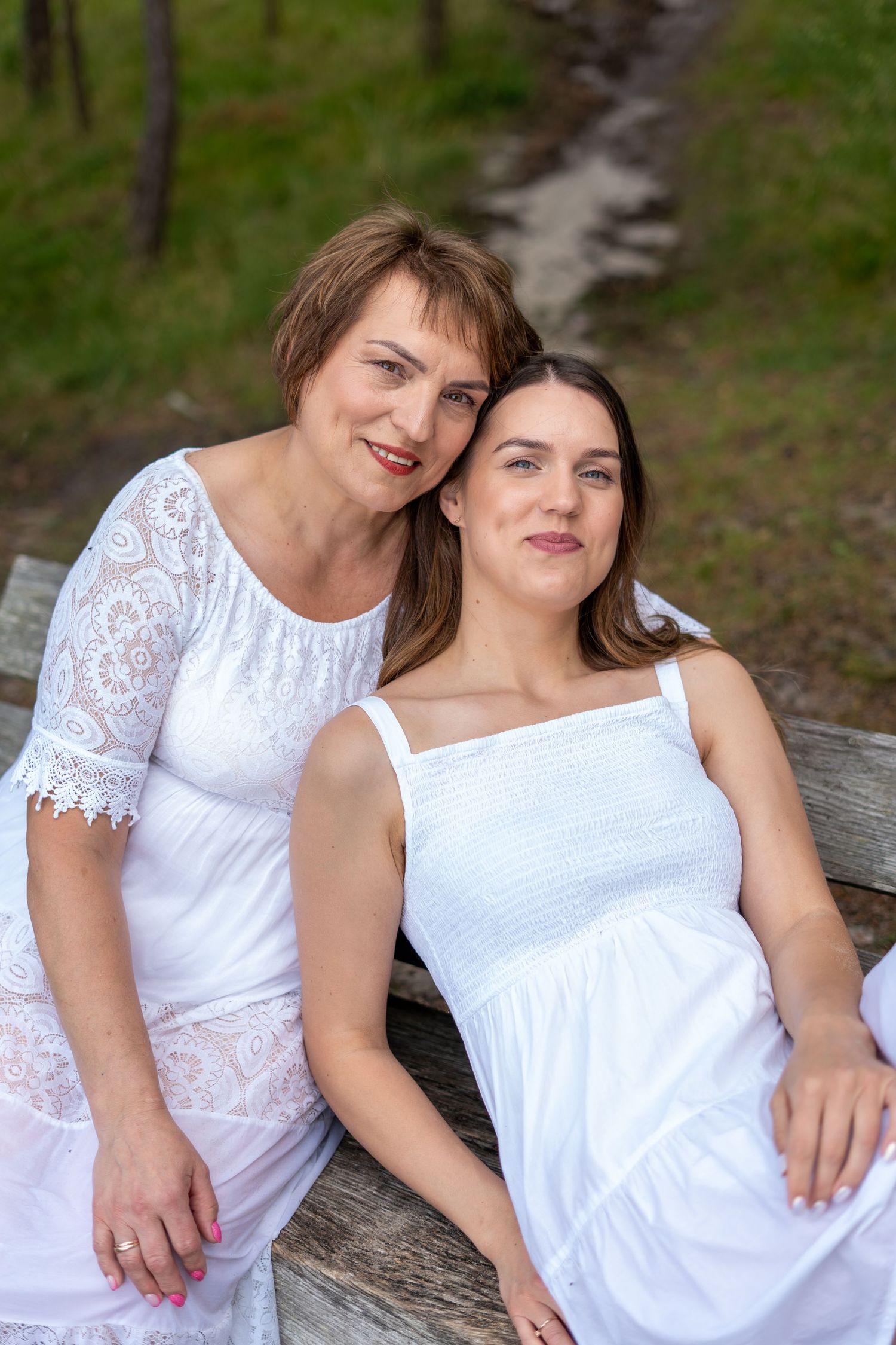 Two women in white dresses sit closely together on a wooden bench in a forest setting.