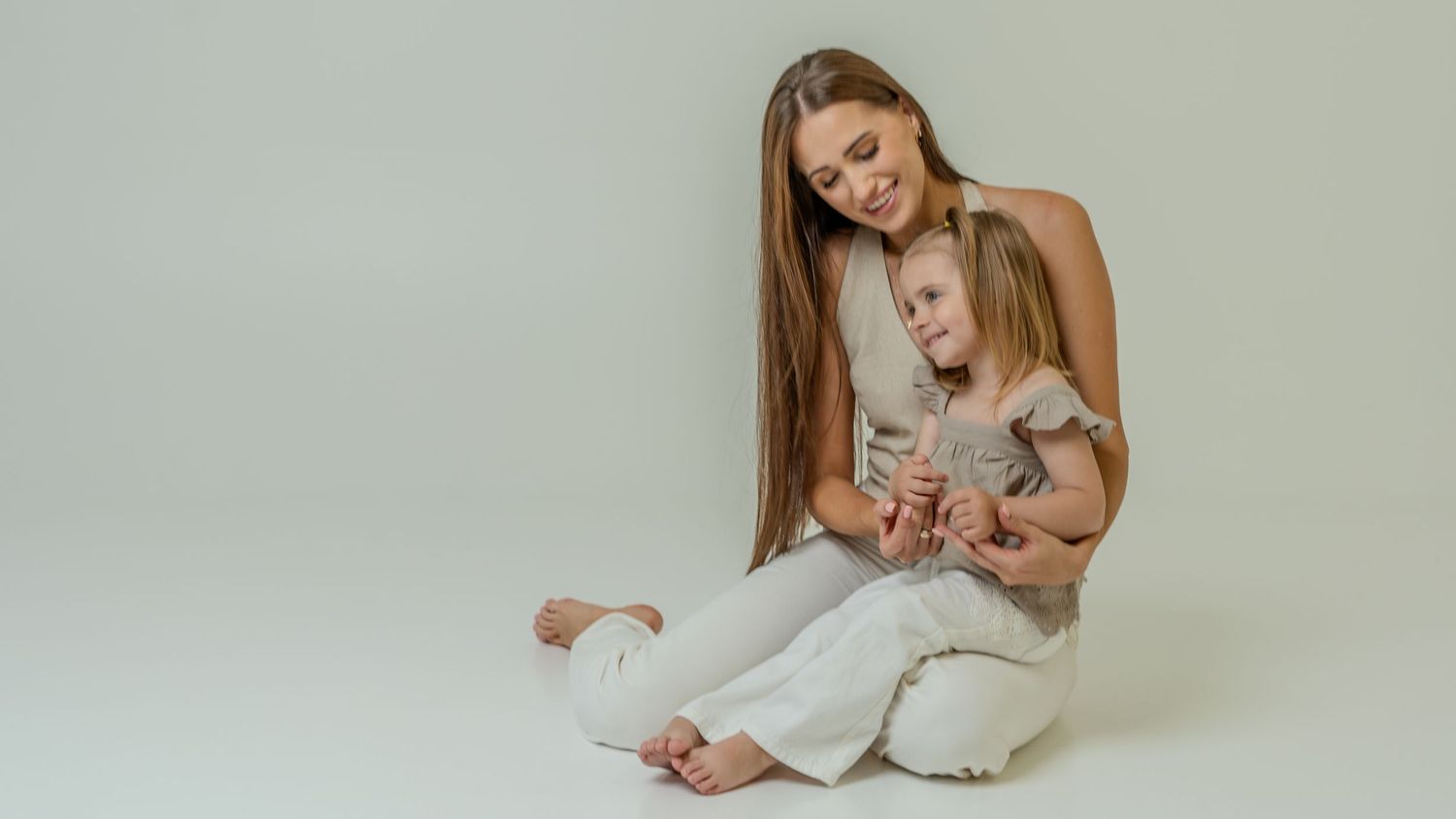Brunette mother with long hair sitting on floor hugging young daughter in light gray outfit on white background.