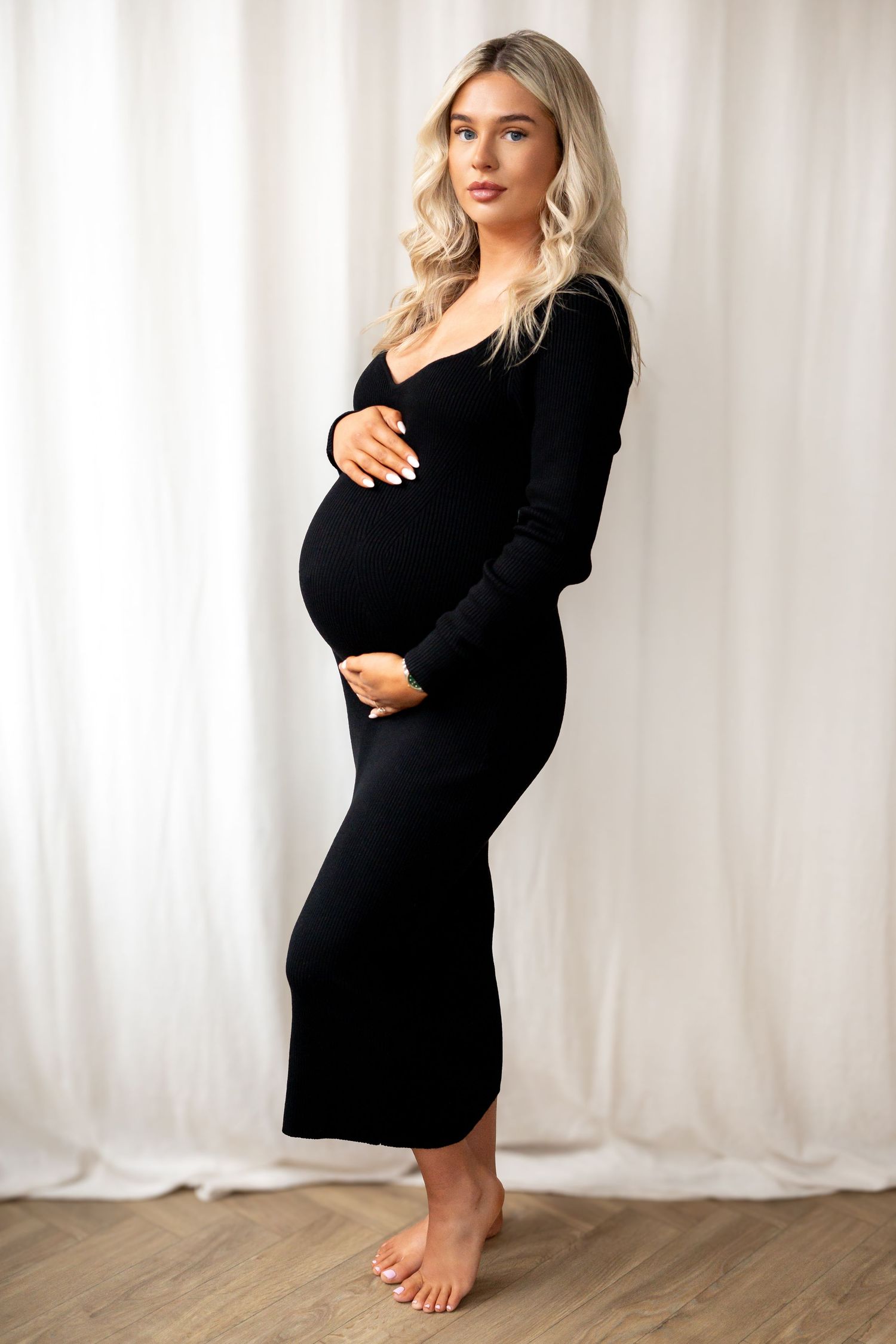 Full length portrait of pregnant woman in black ribbed dress holding bump, barefoot on wood floor.