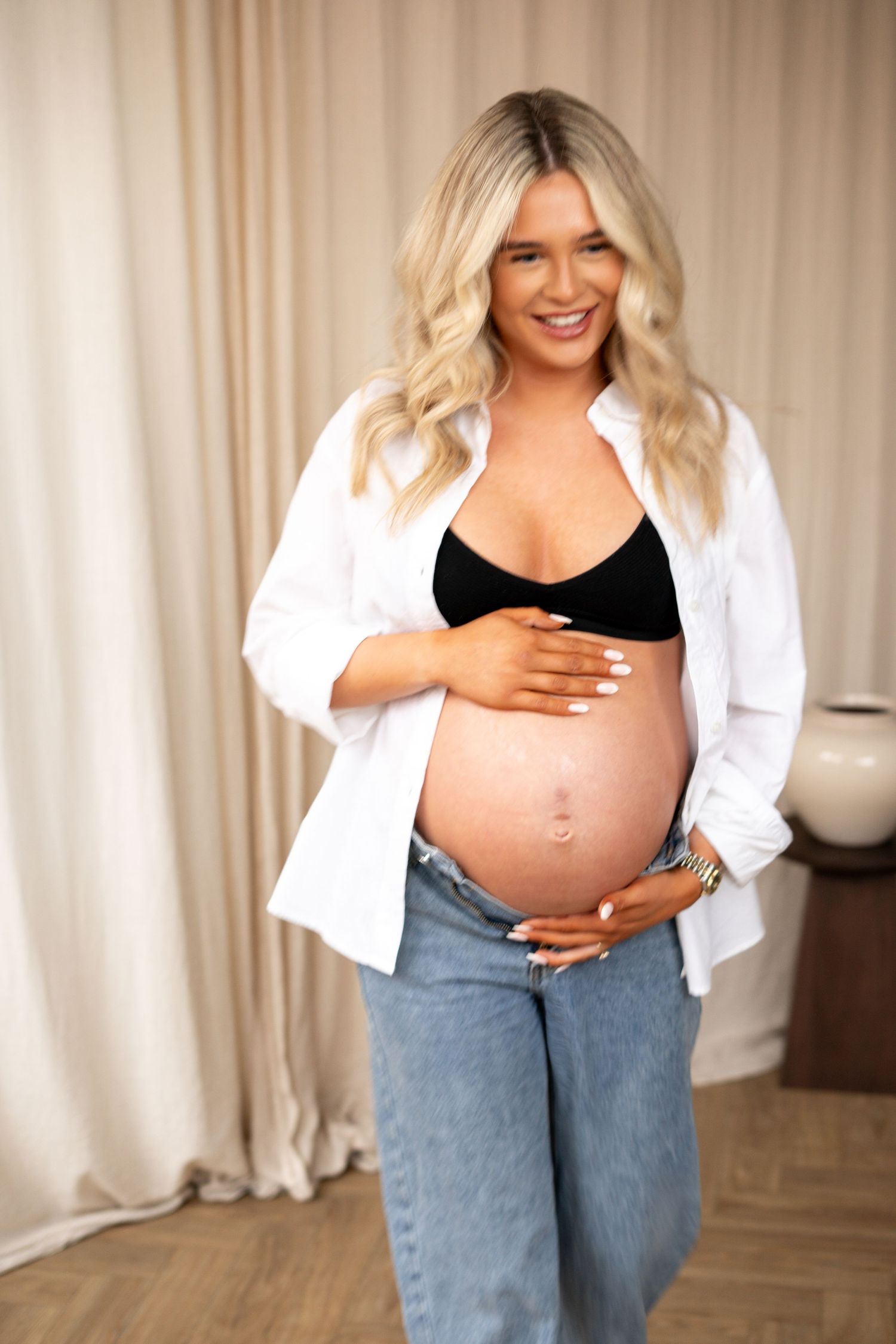 Happy pregnant blonde woman in white shirt and black bralette smiling while holding bump.