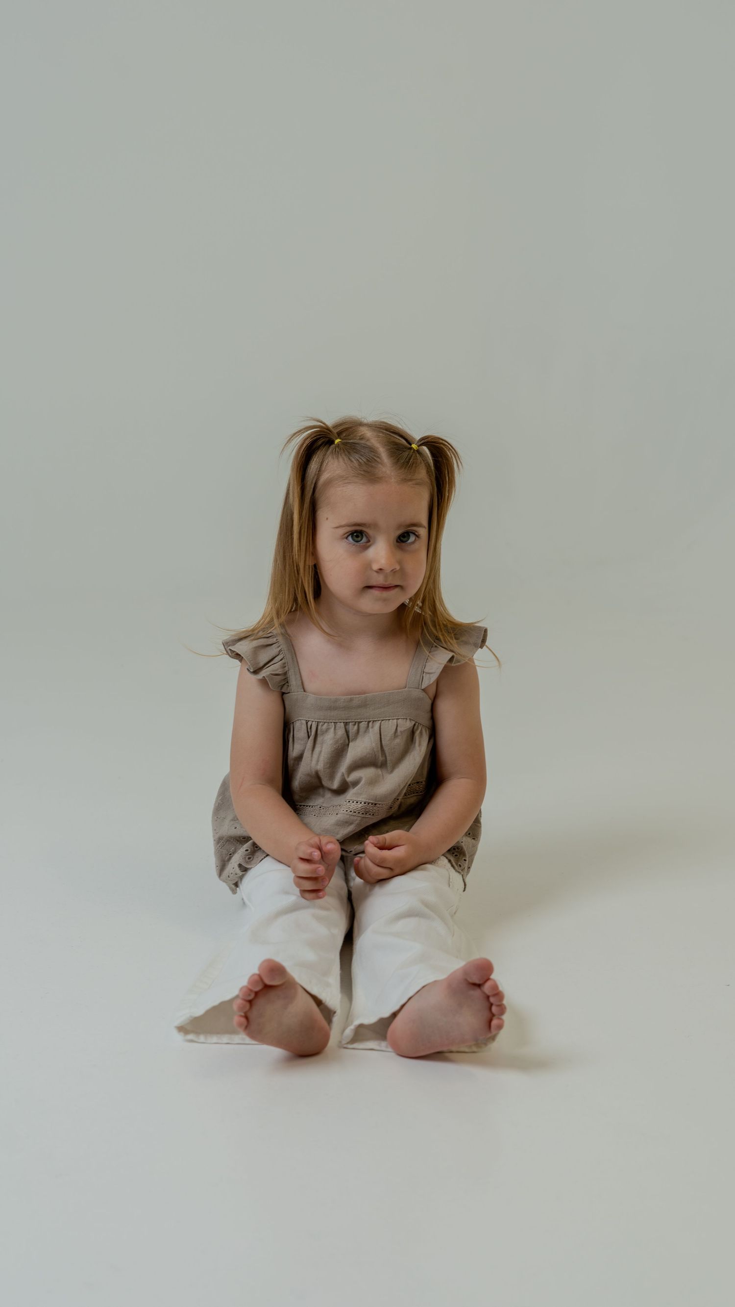 Young blonde girl with pigtails sitting cross-legged barefoot in white pants and gray top on white background.