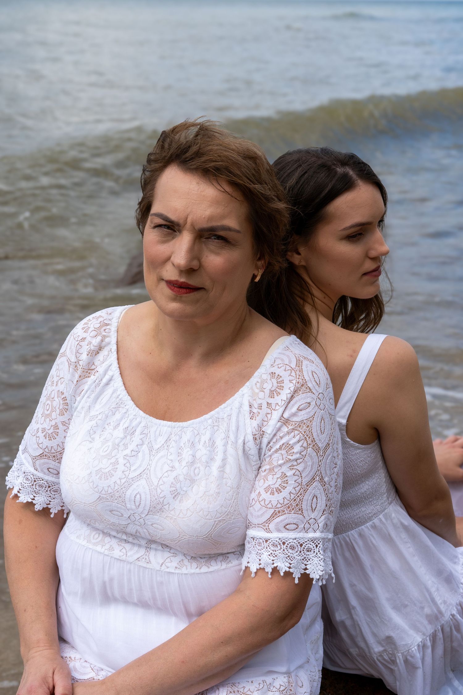 Two women in white dresses stand back-to-back on a beach with ocean waves behind them.