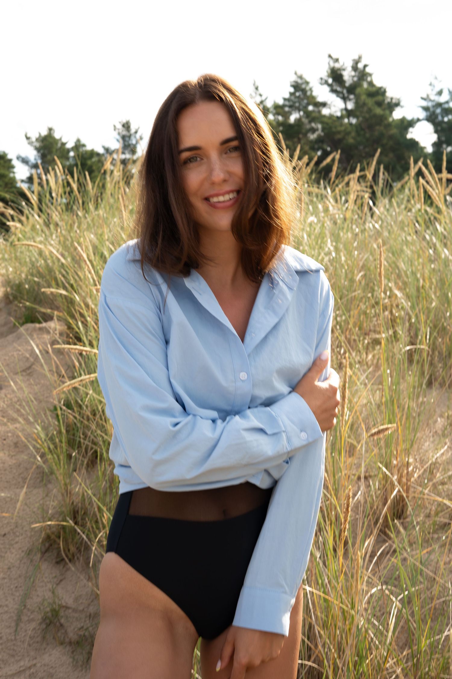 Smiling brunette woman in blue shirt and black bikini bottoms poses in coastal grass dunes.