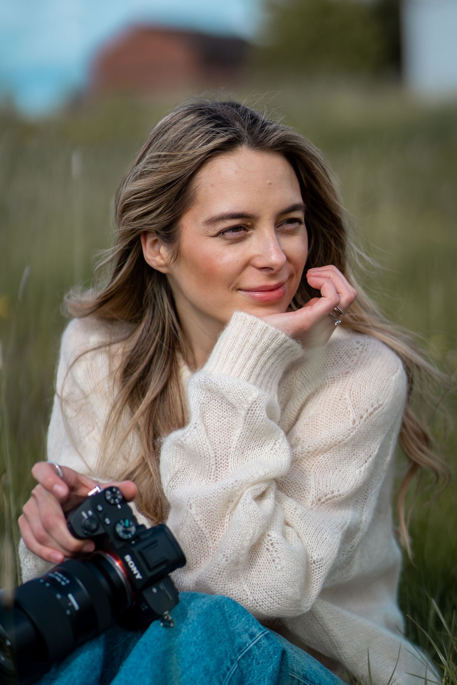 A smiling woman with long blonde hair sits in tall grass holding a camera, wearing a cream knit sweater and jeans.