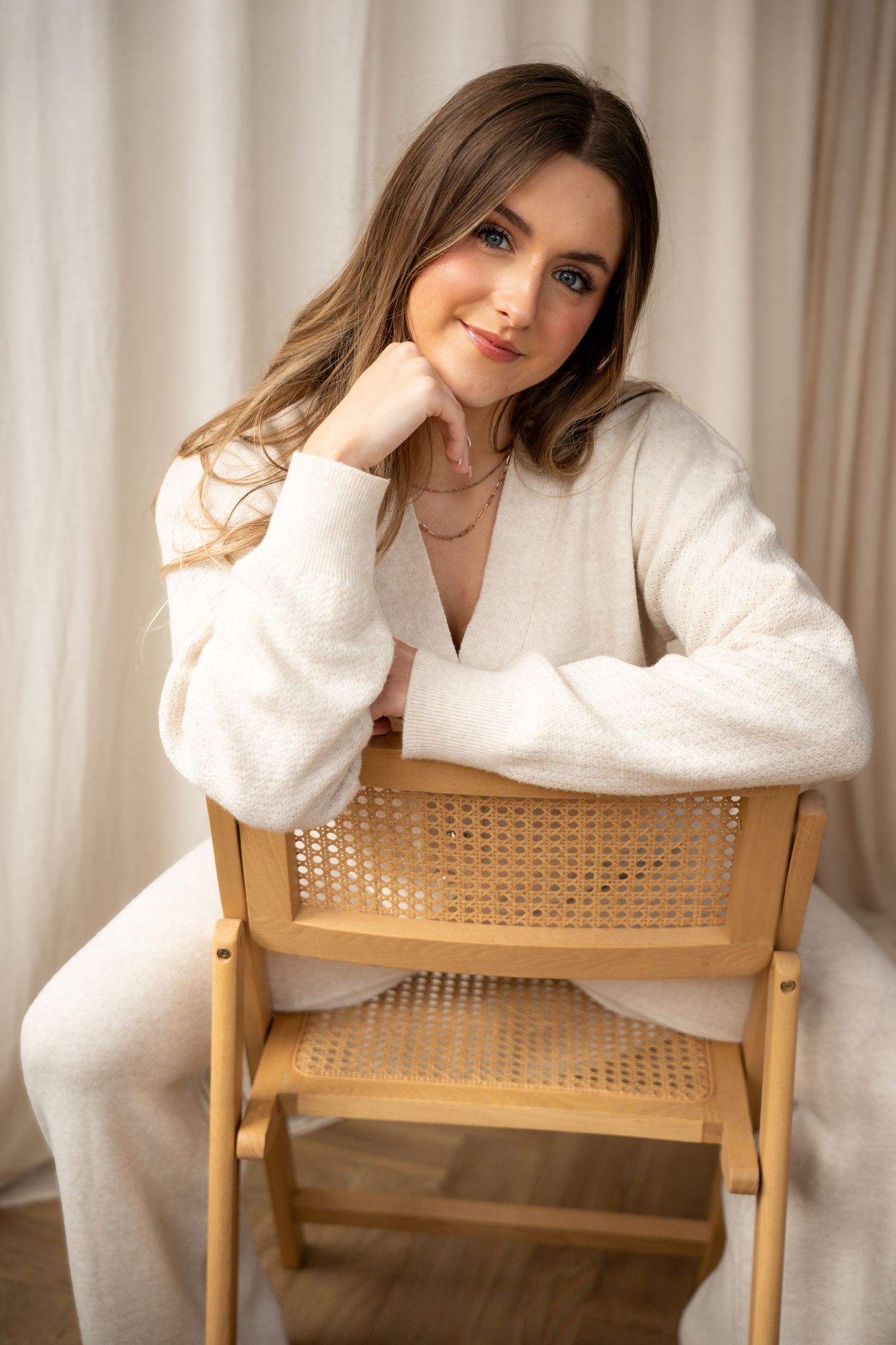 Elegant brunette woman in white cardigan sits on a wooden rattan chair with a gentle smile.