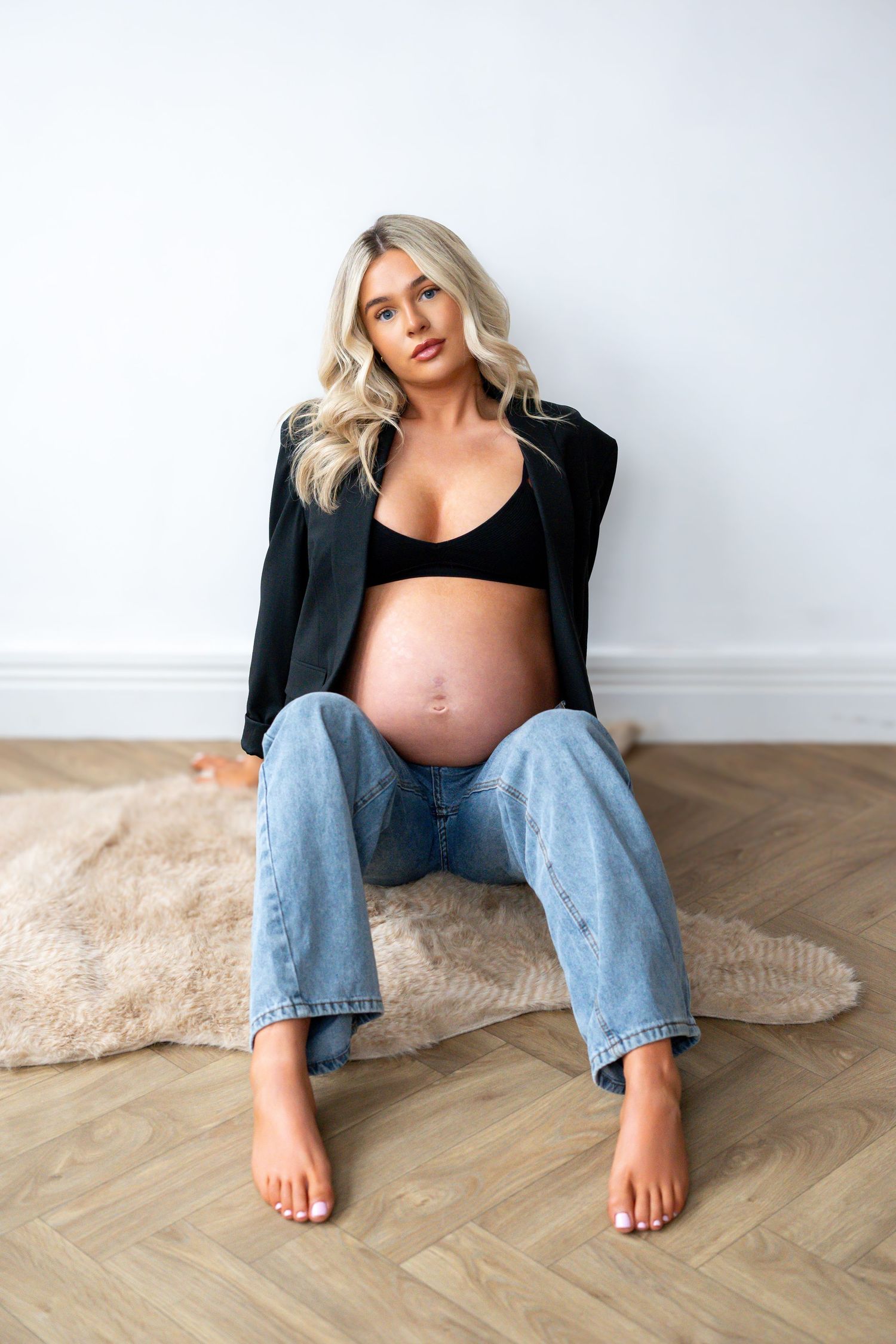 Barefoot pregnant woman sitting on rug in black blazer and jeans with exposed baby bump.