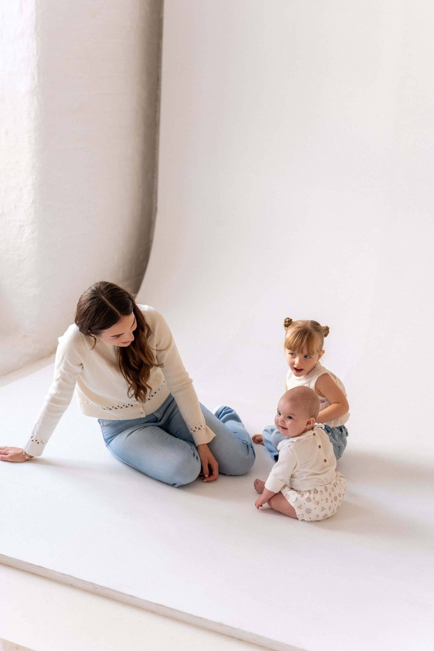 A young mother sits on a white studio floor smiling at her two small children playing beside her.