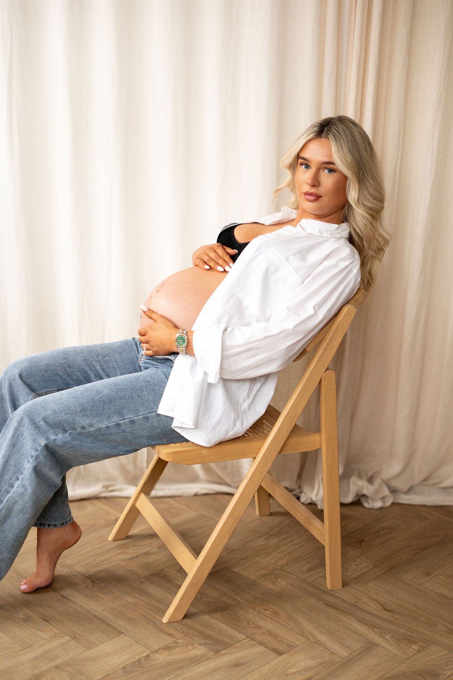 Pregnant woman in white shirt and jeans sitting on wooden chair cradling baby bump in bright studio.