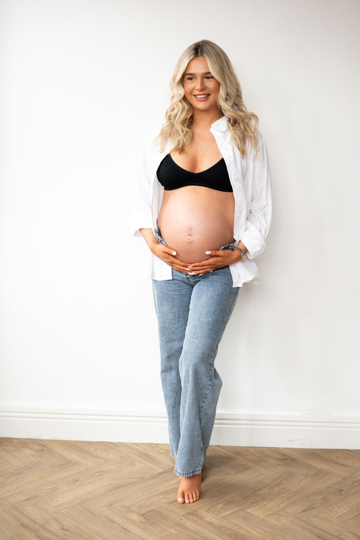 Smiling pregnant woman in white shirt and jeans standing against white wall holding bump.