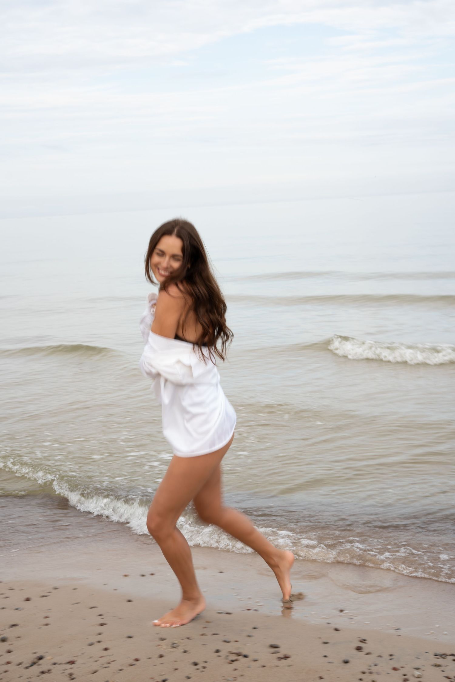 Happy brunette woman in white off-shoulder shirt walking barefoot along ocean shoreline.