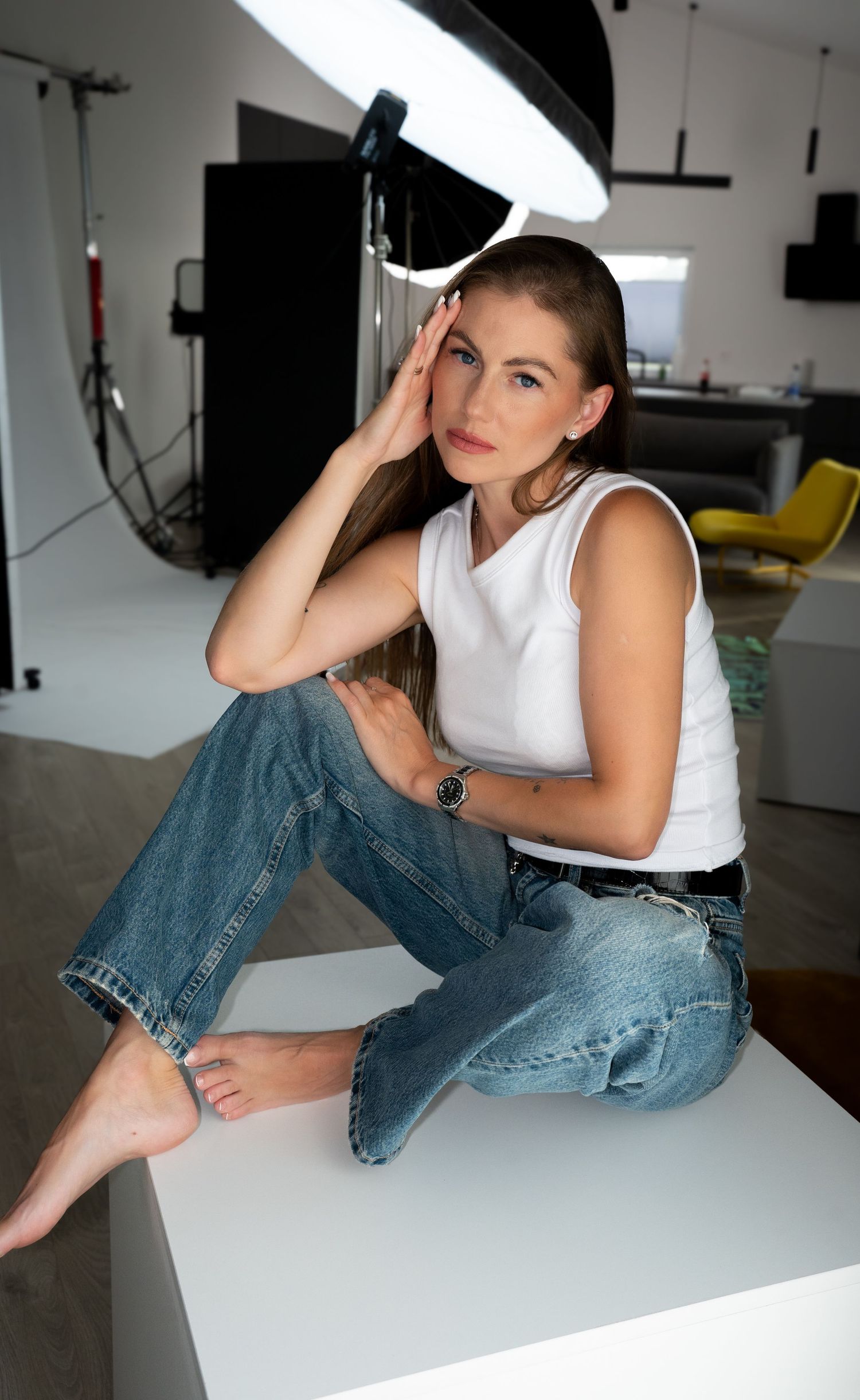 Woman in white sleeveless top and blue jeans sits barefoot on white surface in professional photo studio.