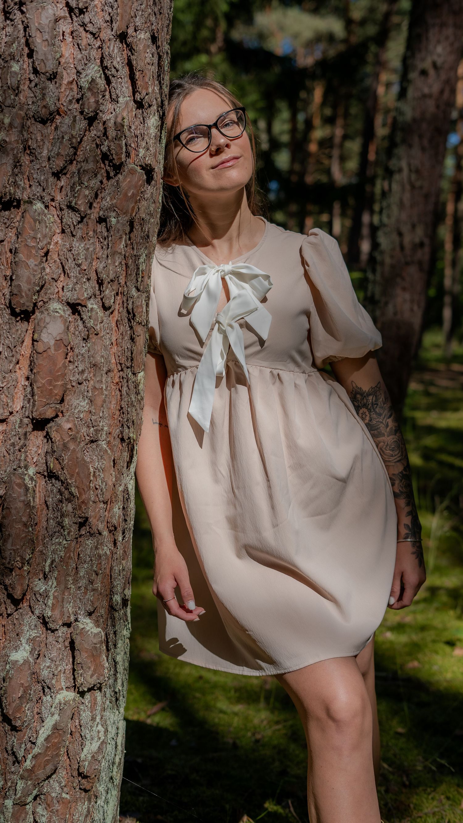Woman with glasses and tattoos in a beige bow dress leans against a pine tree in a forest.
