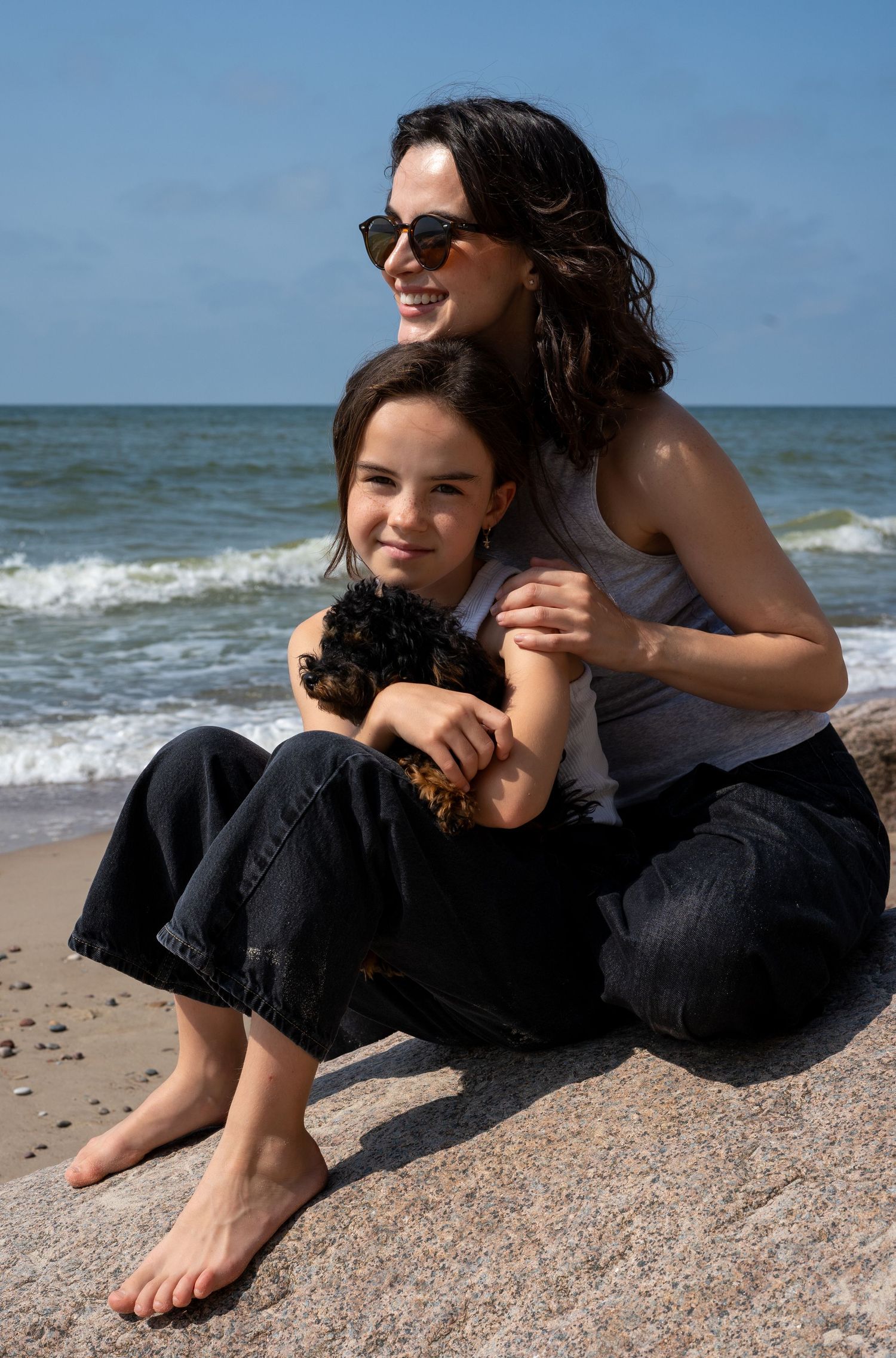 A woman in sunglasses and a girl holding a puppy sit together on a rock by the ocean.