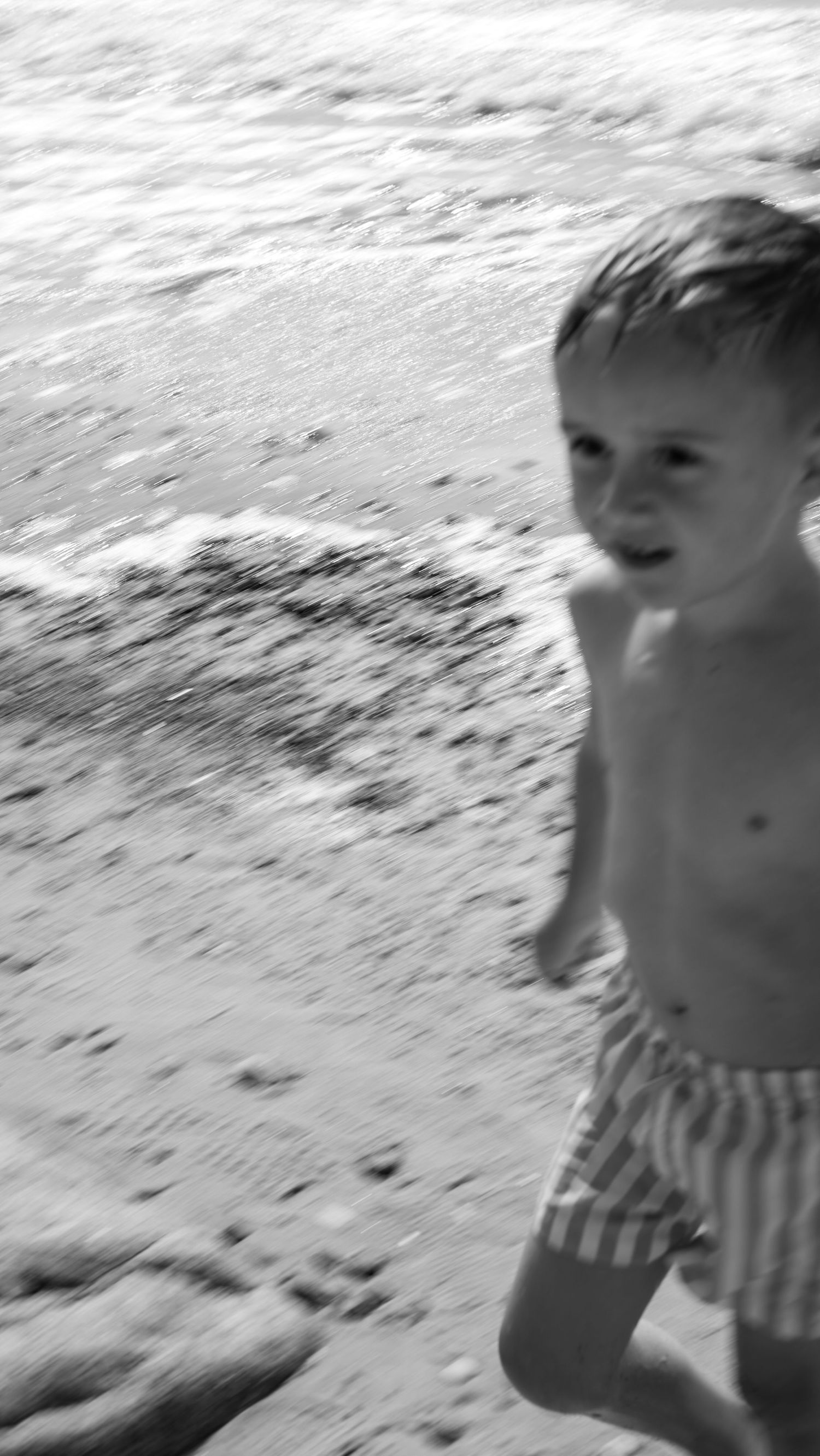 Black and white photo of young boy in striped swim shorts running on sandy beach by waves.