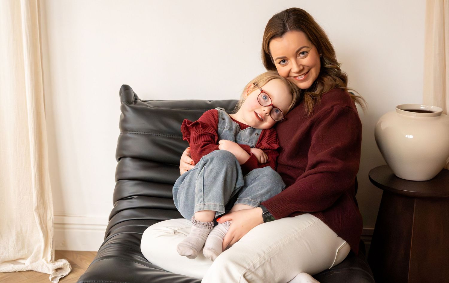 A woman and young girl with glasses cuddle together smiling on a black leather chair.