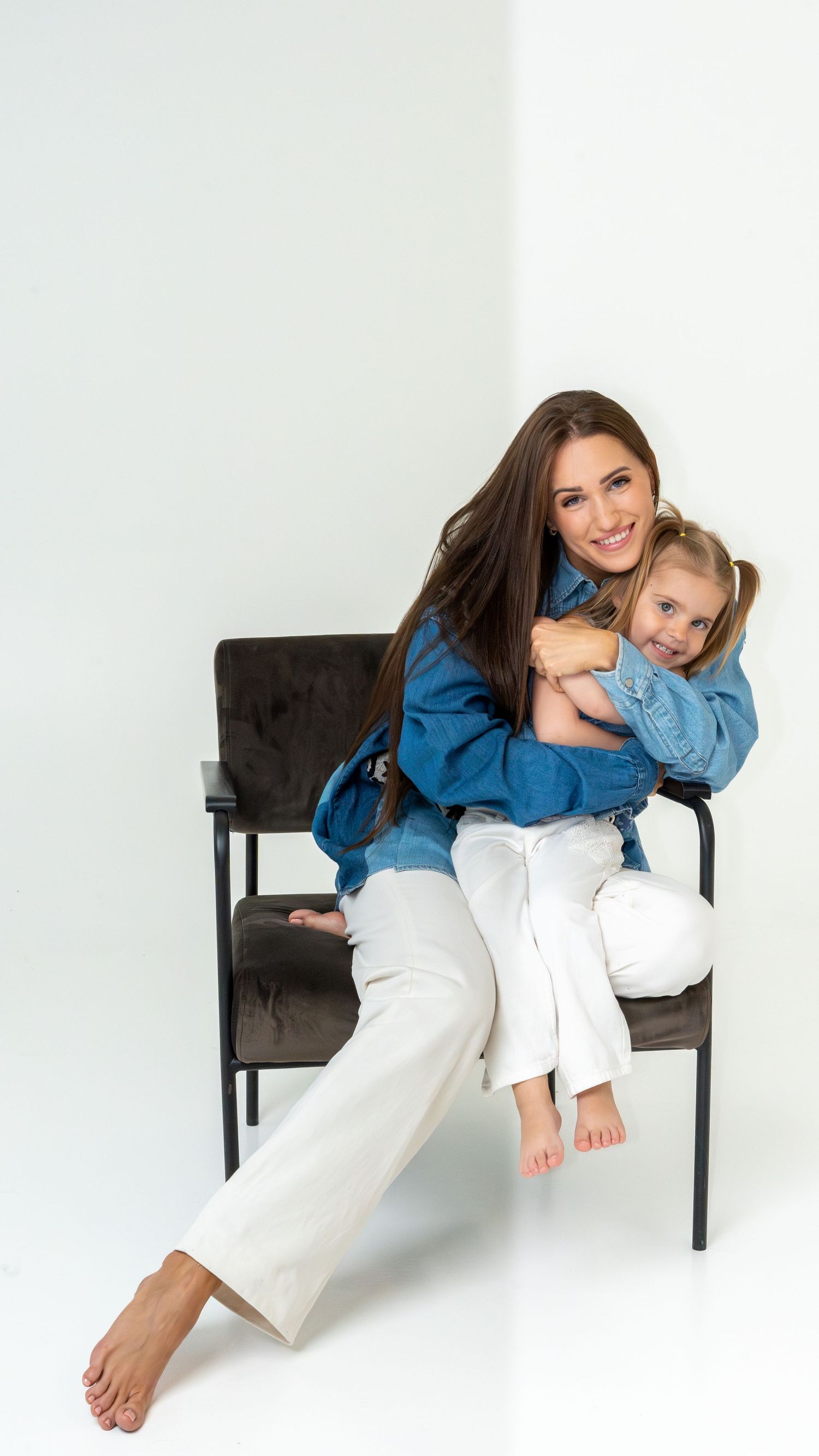 Smiling brunette mother in denim jacket hugging laughing young daughter on chair in white studio.