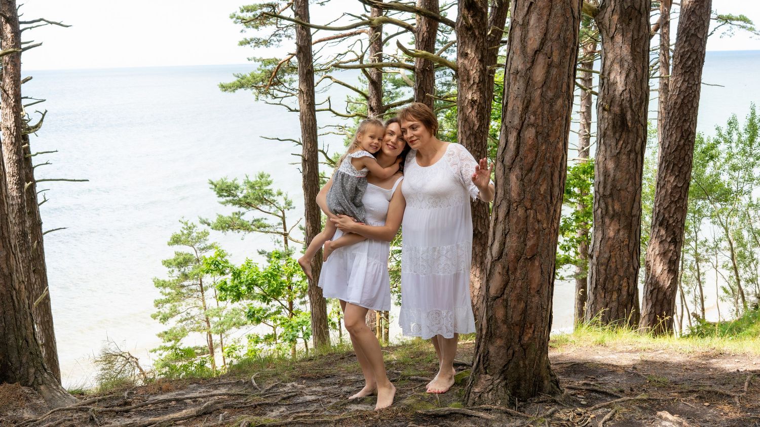 Two women and a young child pose happily among pine trees near a scenic waterfront.