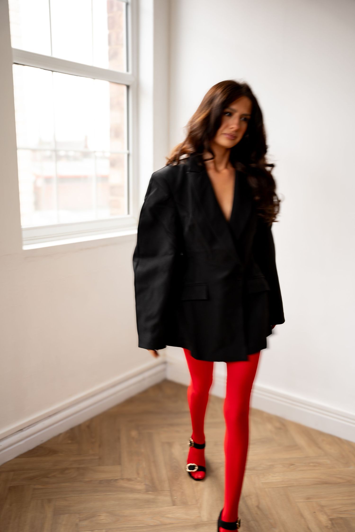 Motion blur shot of brunette woman in black blazer and red tights walking near large window.