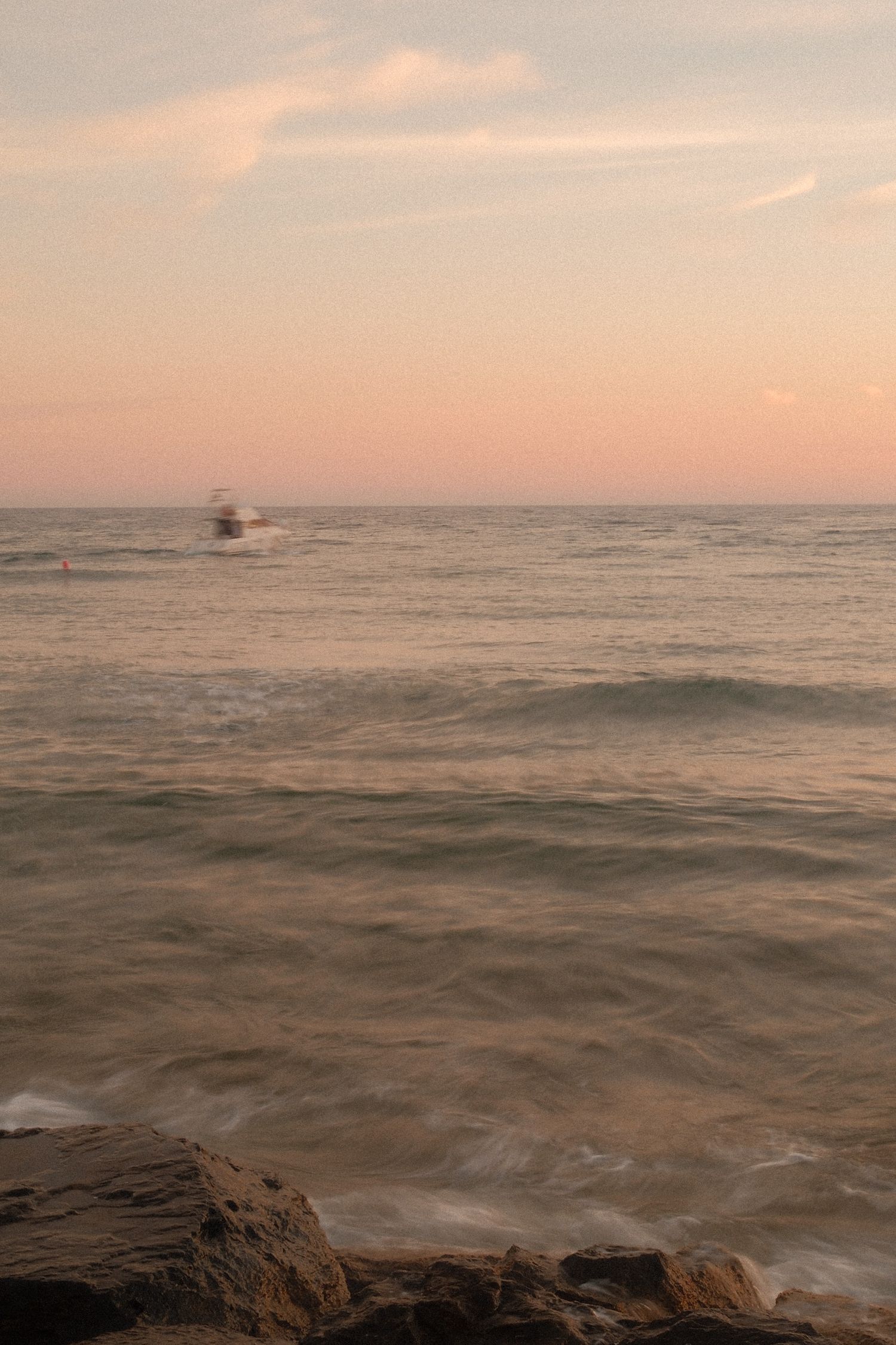 Peaceful ocean sunset with gentle waves, rocky shore, and a boat on the horizon.