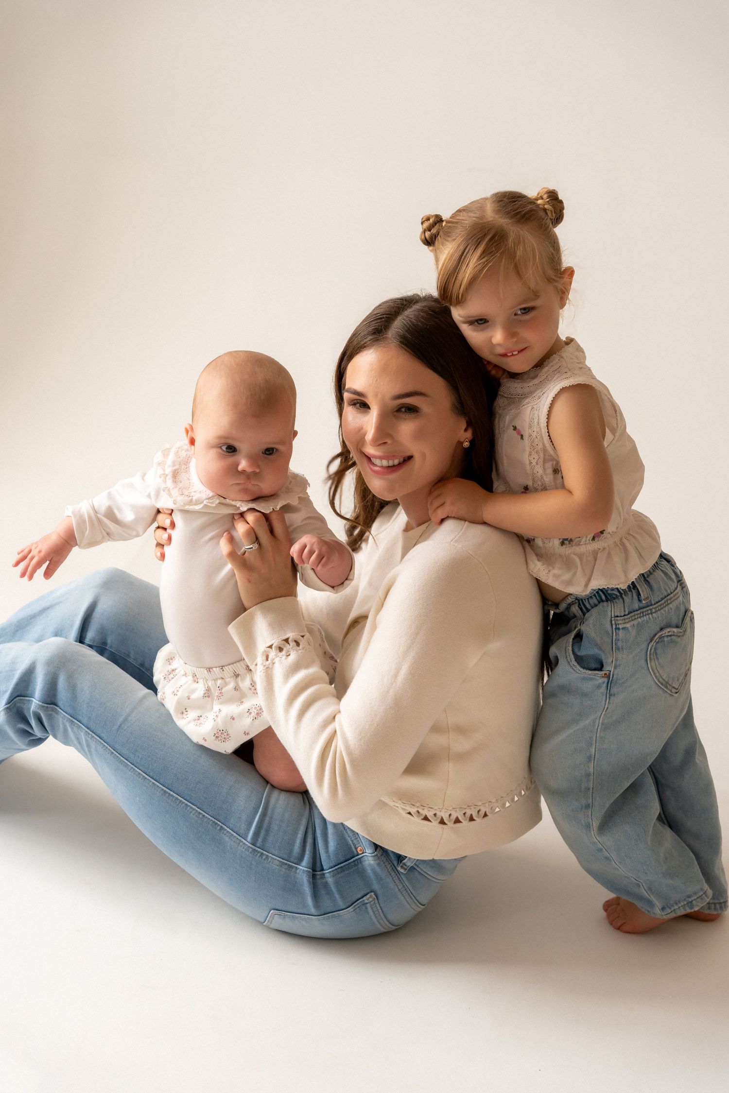 A smiling mother in white sweater and jeans poses with her baby and toddler daughter in a studio photo.